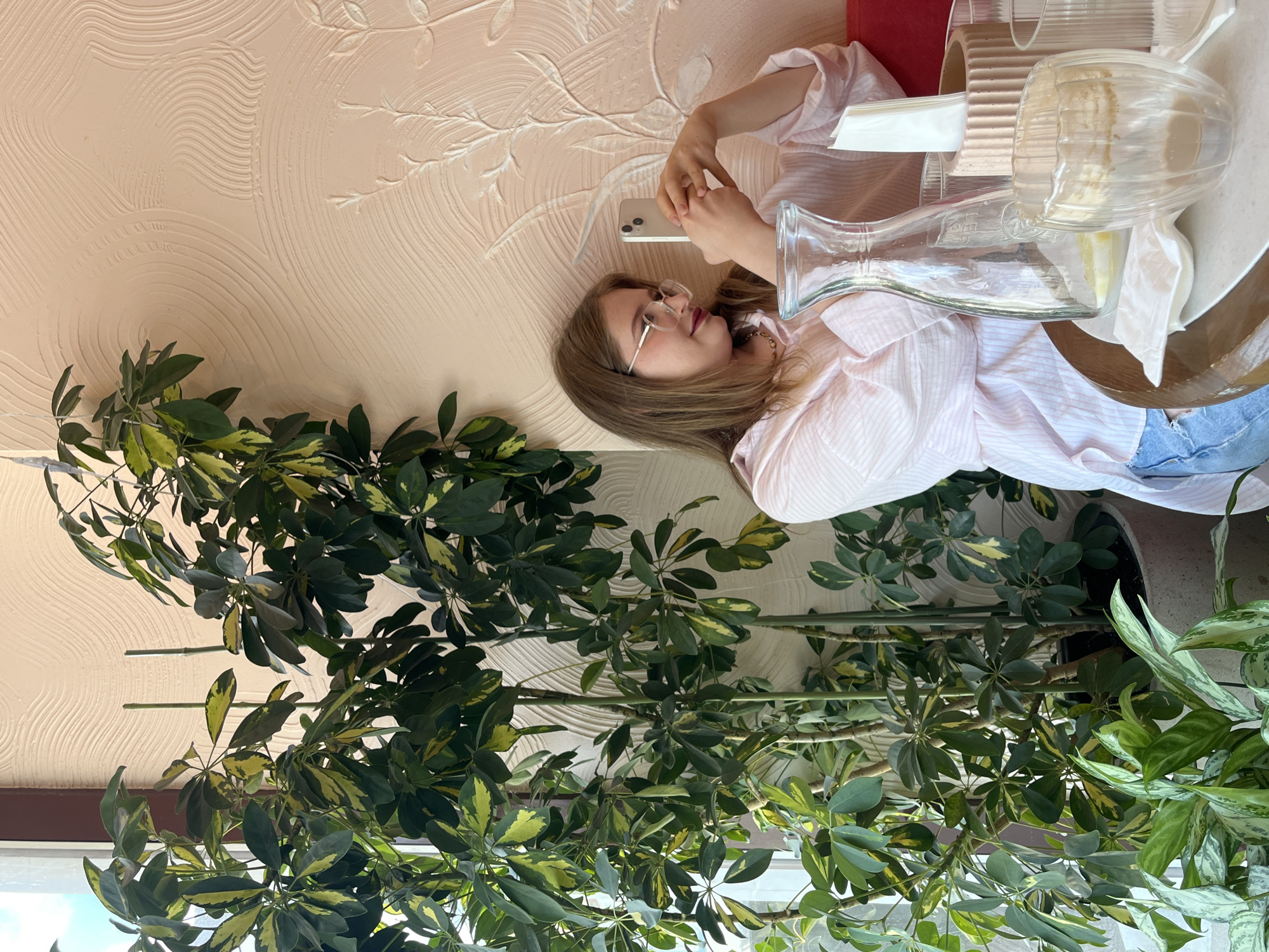 A young woman with light brown hair and glasses is sitting at a table with a large potted plant to her left. She is wearing a pink striped shirt and blue jeans. She is holding a white smartphone in her hands and appears to be taking a picture or recording a video. On the table in front of her are a glass pitcher with some liquid and lemon slices, a glass cup with some residual liquid, a napkin holder with napkins, and other drinking glasses. The wall behind her has a textured pattern. The overall scene suggests a casual setting, possibly a cafe or restaurant, in an unknown city location.