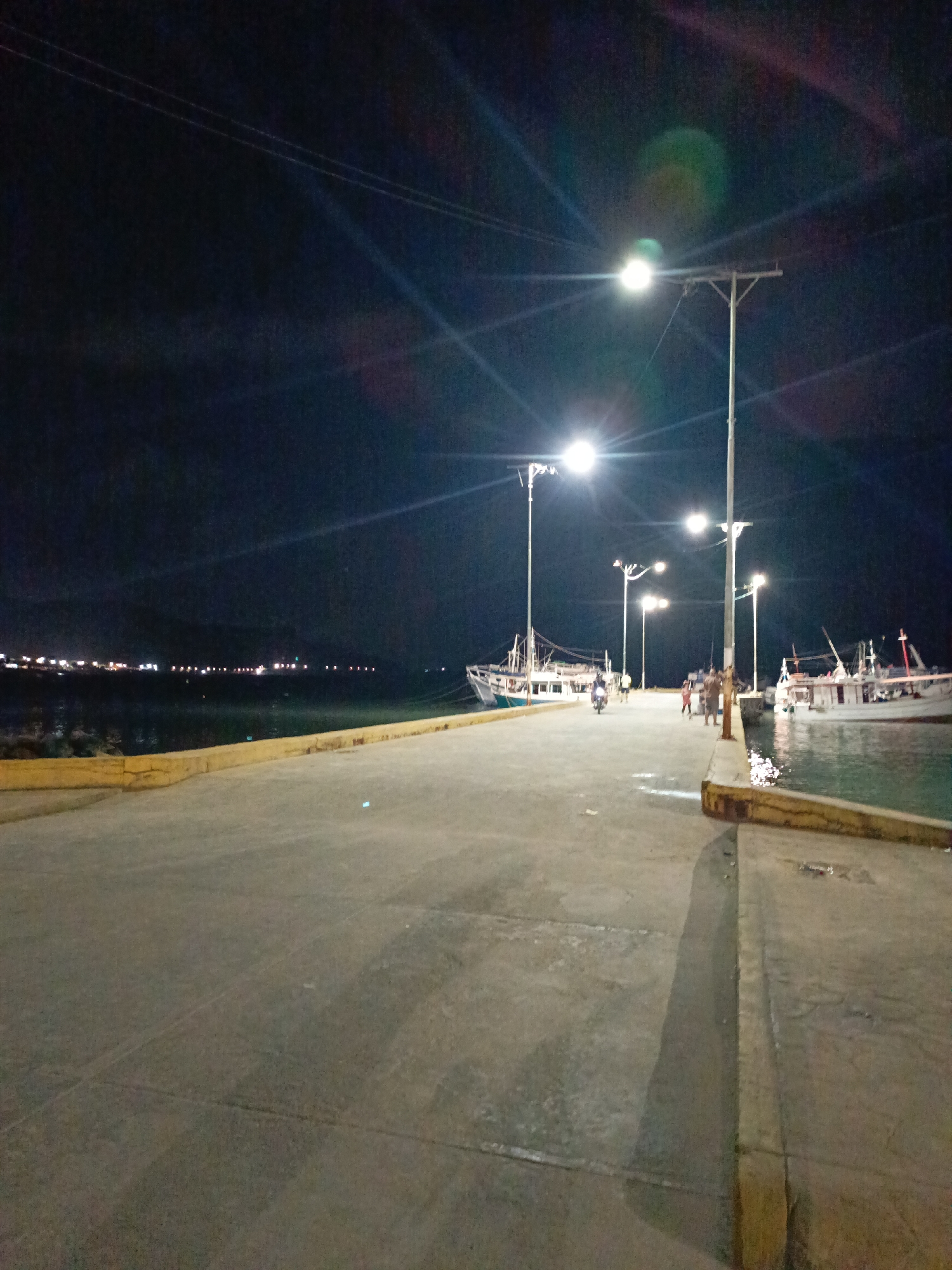 The image depicts a nighttime scene in Juan Griego, Venezuela. A concrete pier stretches into the water. Streetlights illuminate the pier and reflect off the surface of the water. Several boats are docked along the pier's edge. Some individuals are present, with a motorbike and pedestrians visible. In the distance, lights from the town line the horizon across the water. The sky is dark.