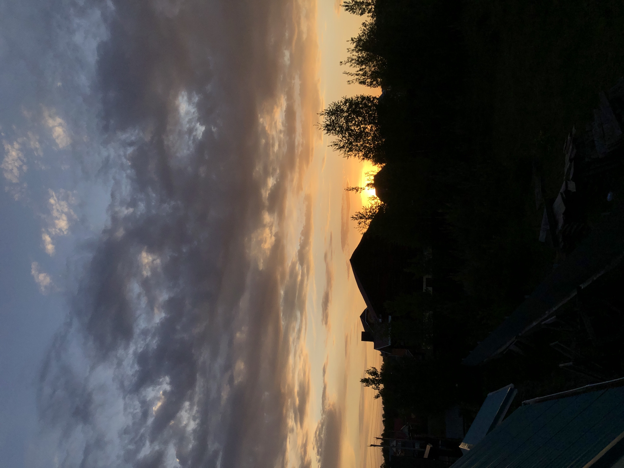 The image captures a sunset in Noviy, Russia. The sky is filled with clouds ranging in color from dark gray to light orange. The sun is low on the horizon and appears to be partially obscured by trees and a building. The silhouette of a house is visible against the bright sky. The foreground is dark, with the roofs of other buildings and vegetation visible.