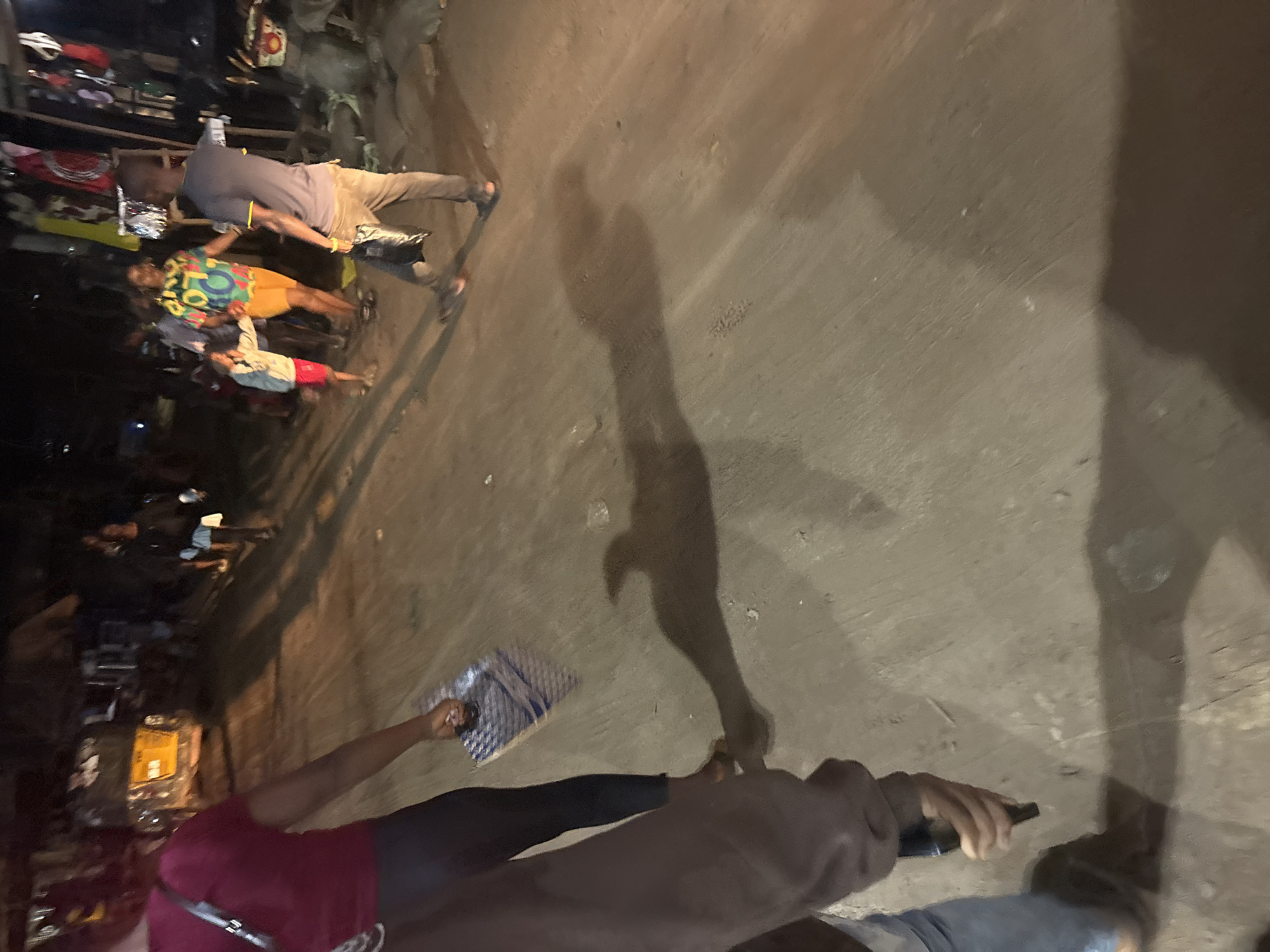 In Nkpor, Nigeria, at nighttime, a group of people are gathered on a dirt road. A woman in a maroon shirt and black leggings, in the foreground to the left, holds a blue and white patterned object in her hand. To her right, a man in a dark gray t-shirt and tan pants, with a black bag in his hand, walks away from the camera, casting a long shadow. Further in the background, a woman in a colorful, patterned shirt and orange shorts stands with a young person in a light gray jacket and red shorts. Behind them, other individuals and market stalls are visible, illuminated by artificial light. A yellow vehicle is parked to the far left. The overall scene depicts a lively street atmosphere at night.