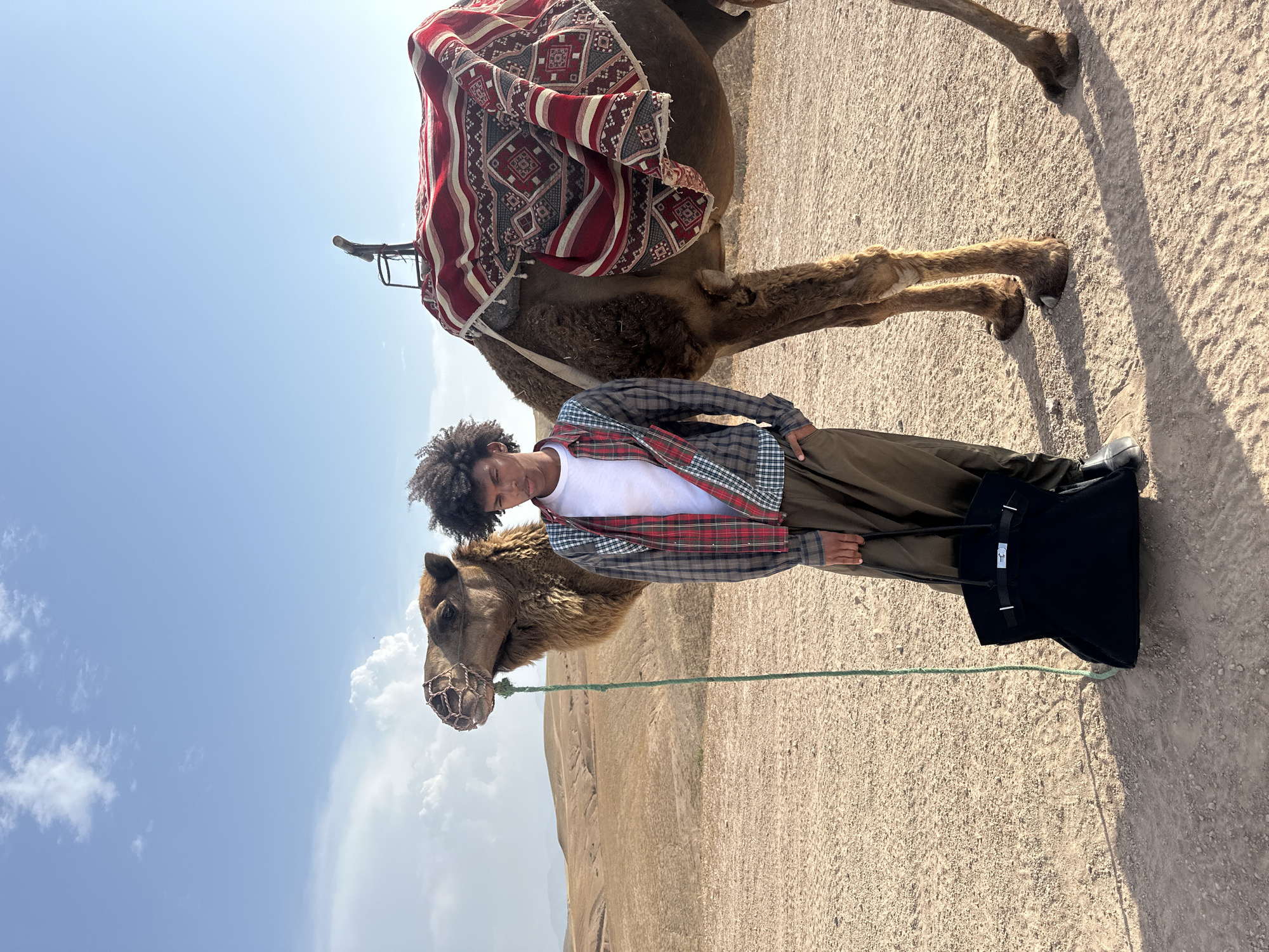 The image captures a scene in Sidi Badhaj, Morocco. A young man stands beside a camel, which is adorned with a colorful saddle blanket. The man is wearing a white t-shirt, a plaid shirt, and dark trousers. He holds the camel's rope. The camel has a muzzle. A black bag sits on the ground near the man's feet. The background is a desert landscape under a partly cloudy sky.
