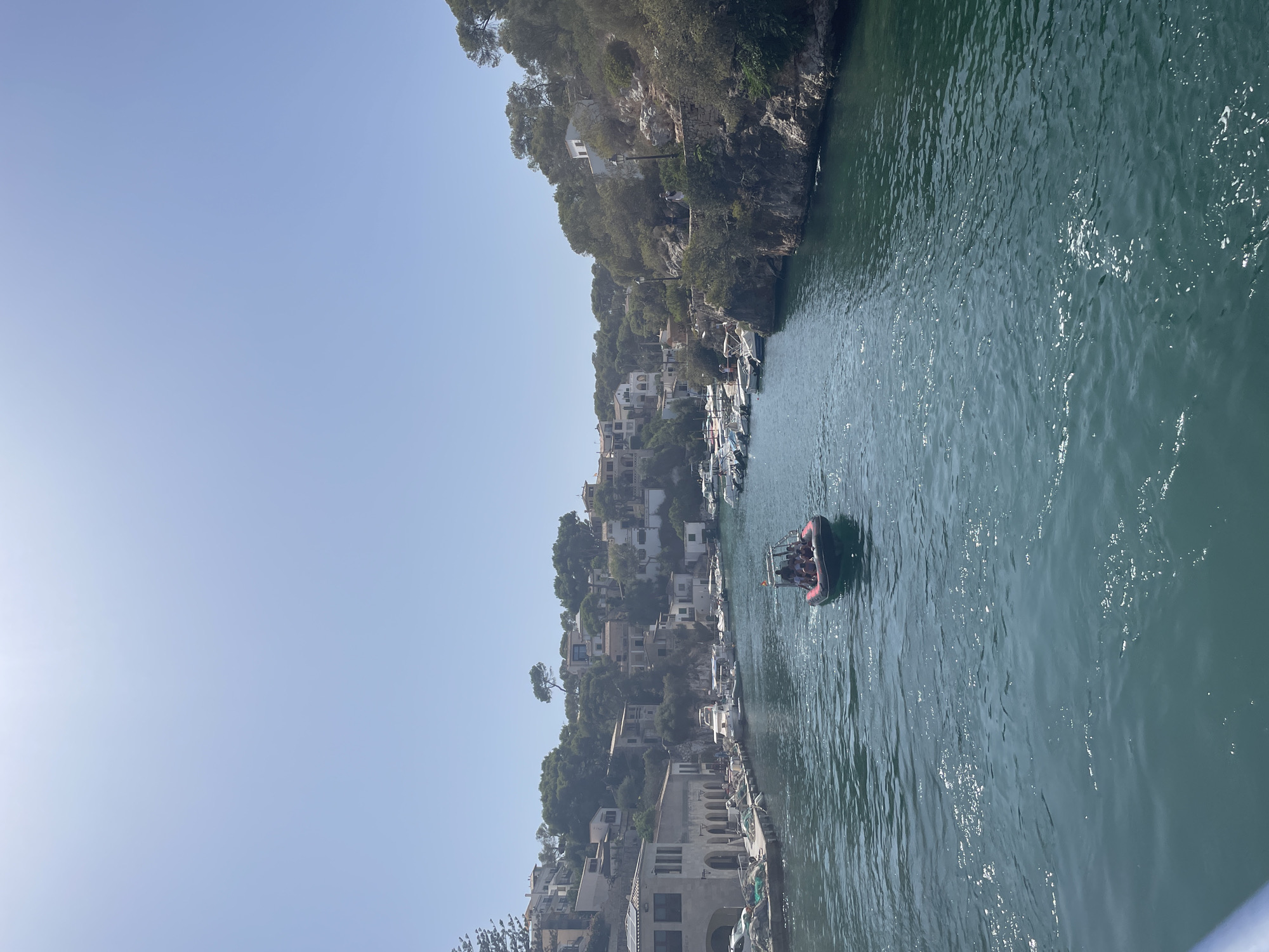 A view from a boat shows a tranquil inlet in Cala Figuera, Santanyí, Spain. The water, a deep blue-green, reflects the bright sunlight, with small waves creating shimmering patterns. On the left, a row of traditional white and stone buildings lines the waterfront, with moored boats and fishing nets visible on the dock. The buildings are set against a backdrop of lush green hills dotted with more houses. On the right, the inlet is bordered by rocky, tree-covered cliffs. A small, red inflatable boat with several people aboard is moving away from the viewer, leaving a wake in the water. The sky is clear and pale blue.