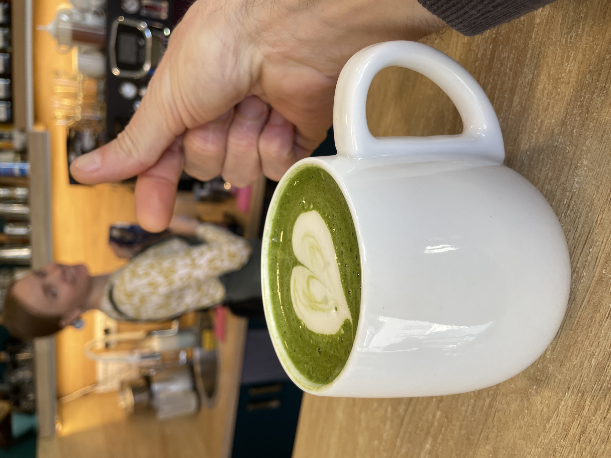 A hand points to a white, round mug filled with matcha latte art on a wooden table. In the background, a blurred woman with short hair stands near a commercial coffee machine. The location cannot be confirmed as Budva, Montenegro from this image alone.