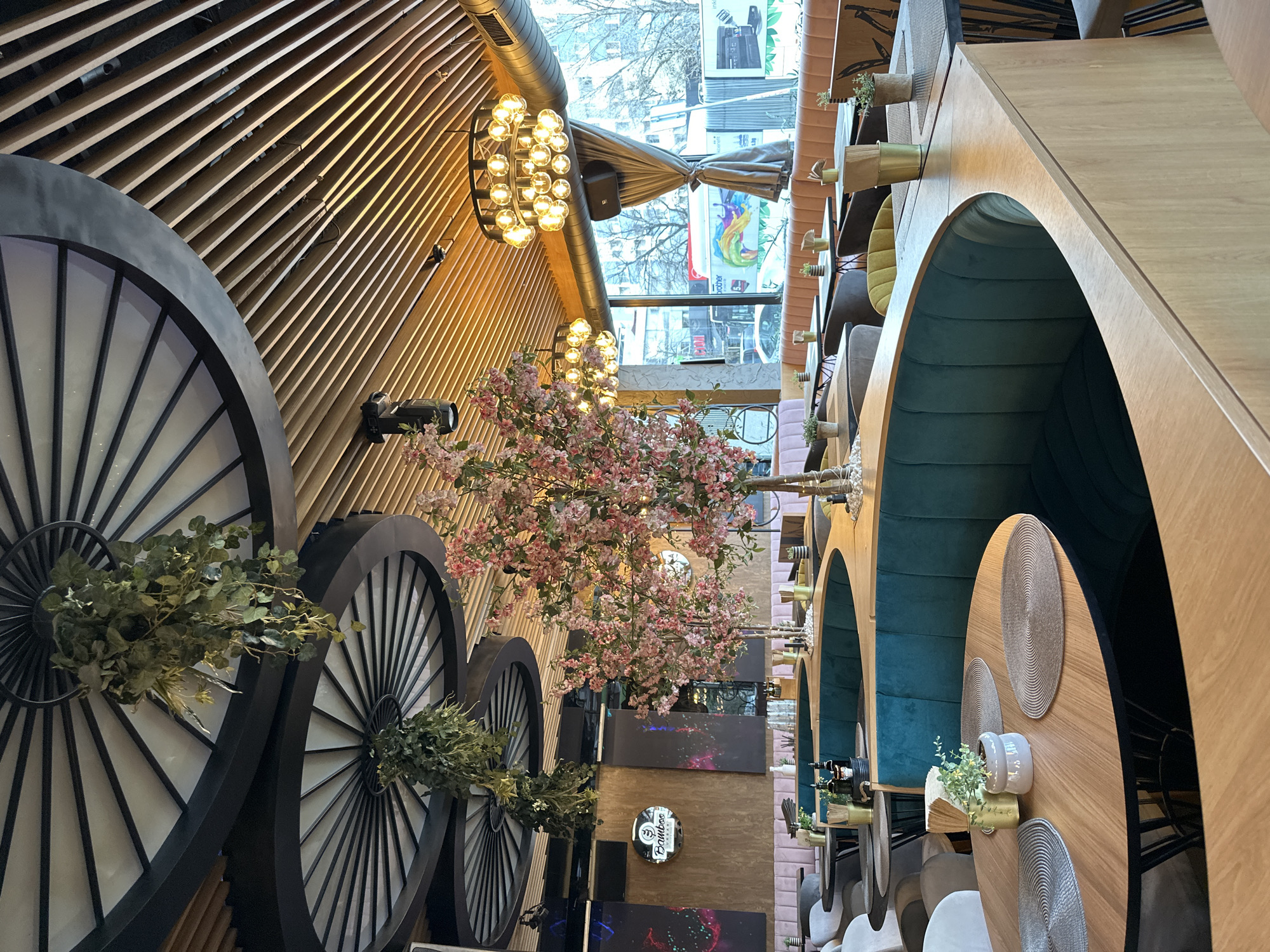 The image depicts the interior of a restaurant in Sofia, Bulgaria. The scene is dominated by a unique design featuring large, decorative wagon wheels mounted on a wall with wooden slat accents. A faux cherry blossom tree extends from the wall, adding a touch of color. Lighting fixtures are a prominent feature, including a modern chandelier near the ceiling. The restaurant has arched booths with blue upholstery along one side, with round wooden tables set for dining. Placemats and small potted plants adorn the tables. Outside the window, glimpses of the city street can be seen. The restaurant seems unoccupied at the time the photograph was taken.
