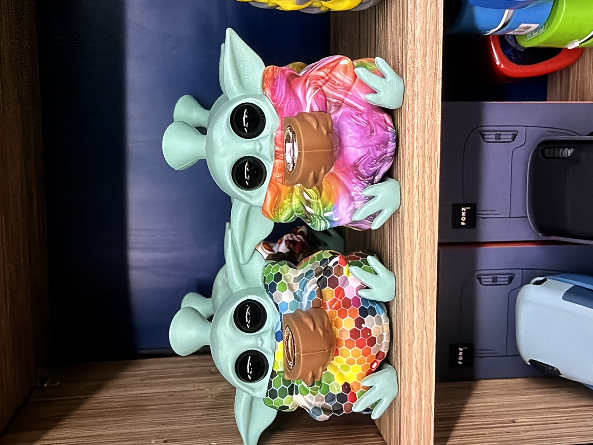 Two Grogu figurines, also known as Baby Yoda, are displayed on a wooden shelf. The top figurine has a colorful swirling pattern on its robe, while the bottom figurine has a colorful hexagonal pattern. Both figurines have large, black eyes and are holding a cylindrical object. The shelf appears to be part of a storage unit. The background features a blue surface and other objects, including what appears to be a green and blue container and boxes with the word "FONZ" printed on them. The location is unknown.