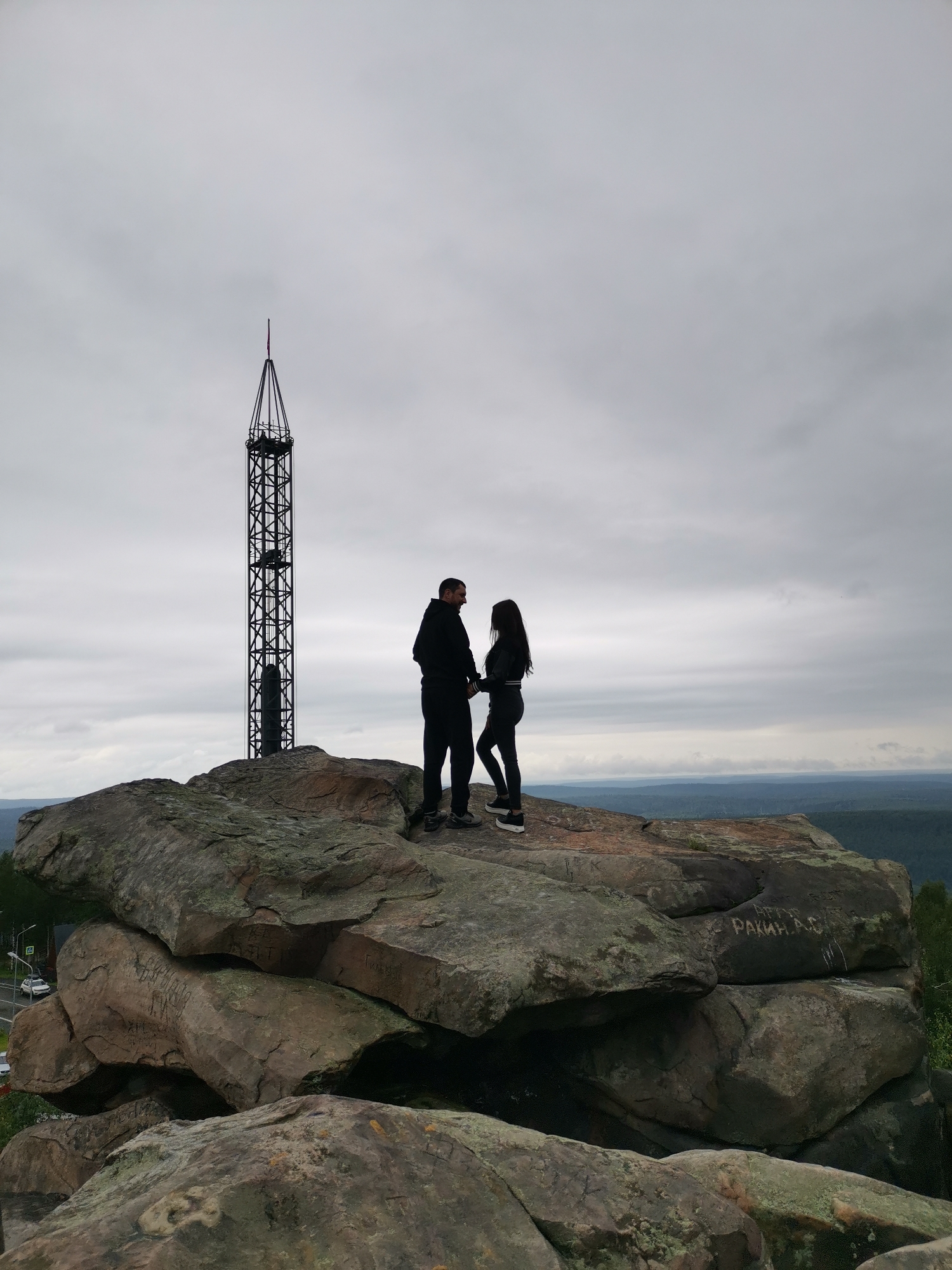 A man and a woman stand on a rocky outcrop, facing each other. The man is on the left, wearing a dark hoodie and pants. The woman is on the right, wearing a dark jacket and pants. Behind them, a tall, metallic tower stands against a cloudy sky. In the distance, a valley filled with trees stretches towards the horizon. The rocky outcrop has visible inscriptions on its surface. The location appears to be the rock formations of Mount Katchkanar, near the city of Gubakha, Russia, known for its television tower and scenic views.