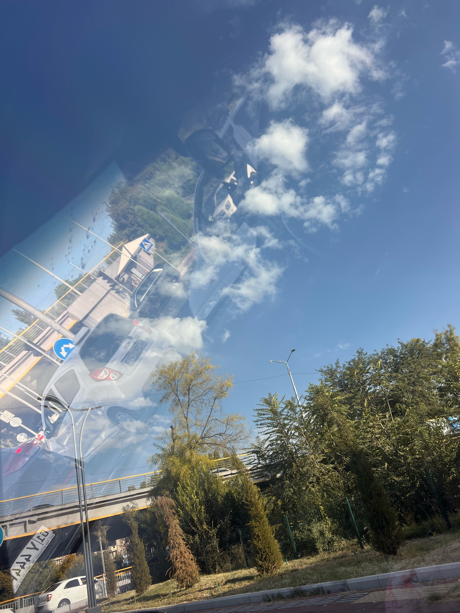 The image is a reflection on a surface, possibly a car windshield or a window, of a city scene with a bright blue sky and scattered white clouds. The scene appears to be taken from a moving vehicle, with the perspective tilted. In the foreground, there are reflections of cars, a street sign, and a pedestrian crossing sign. A white car is visible in the lower left of the image. Behind the reflections, the actual scene shows a bridge or overpass, lush green trees, and a street light. The overall impression is one of urban infrastructure juxtaposed with natural elements under a clear sky.