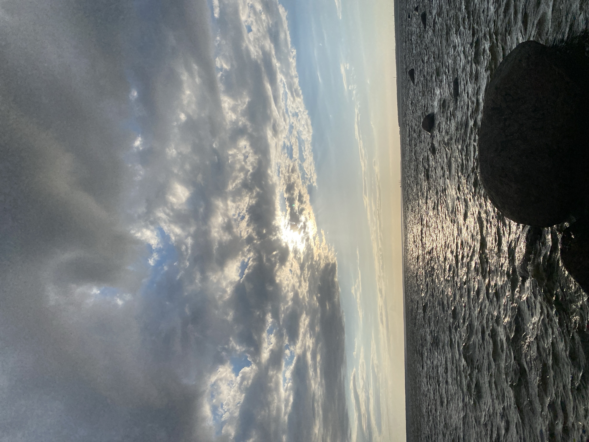 The image depicts a body of water with a rocky shoreline under a cloudy sky. The sun is partially visible through a break in the clouds, casting a shimmering reflection on the water's surface. Several large, dark rocks are scattered in the foreground and mid-ground, some partially submerged in the water. In the distance, a faint line of the horizon suggests a landmass or coastline, with a few indistinct structures visible. The overall scene conveys a sense of natural landscape, likely a coastal area, with overcast weather conditions. There are no people or animals visible in the image, and no apparent actions or interactions are taking place. The setting appears to be a natural environment characterized by water, rocks, and sky.