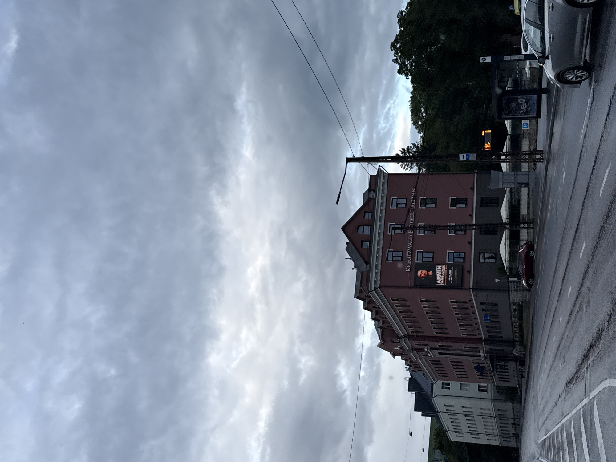 The image depicts a street scene in Tallinn, Estonia, on a cloudy day. The main focus is a reddish-brown building with multiple stories and a pitched roof, identified by signage as the "KREUTZWALD HOTEL TALLINN". A large poster featuring a man's face and text is visible on the building's facade. Across the street, a multi-lane road is wet, suggesting recent rain. White lane markings are present on the road. To the left of the main building, a lighter colored building is partially visible. In the foreground, the wet road surface is prominent, with white pedestrian crossings. A silver sedan is parked on the right side of the road. Further to the right, there is a bus stop shelter with advertisements. Trees are visible in the background. Power lines and a street lamp are also present in the scene.