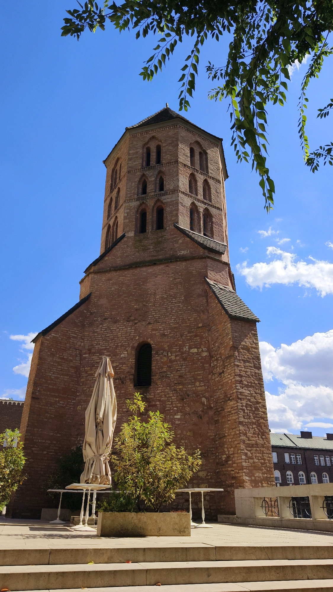 A low-angle shot shows a tall, octagonal brick tower with arched windows and a dark roof, silhouetted against a bright blue sky with scattered white clouds. The tower, which is part of a building resembling a church or historical structure, has a light brown brick facade. In the foreground, a patio area is visible with concrete steps leading up to it. On the patio, there are white tables and chairs, with one table covered by a beige umbrella. A small tree with green foliage grows in a planter box. The scene appears to be a sunny day. The building and tower are located in the city of Szeged, Hungary.