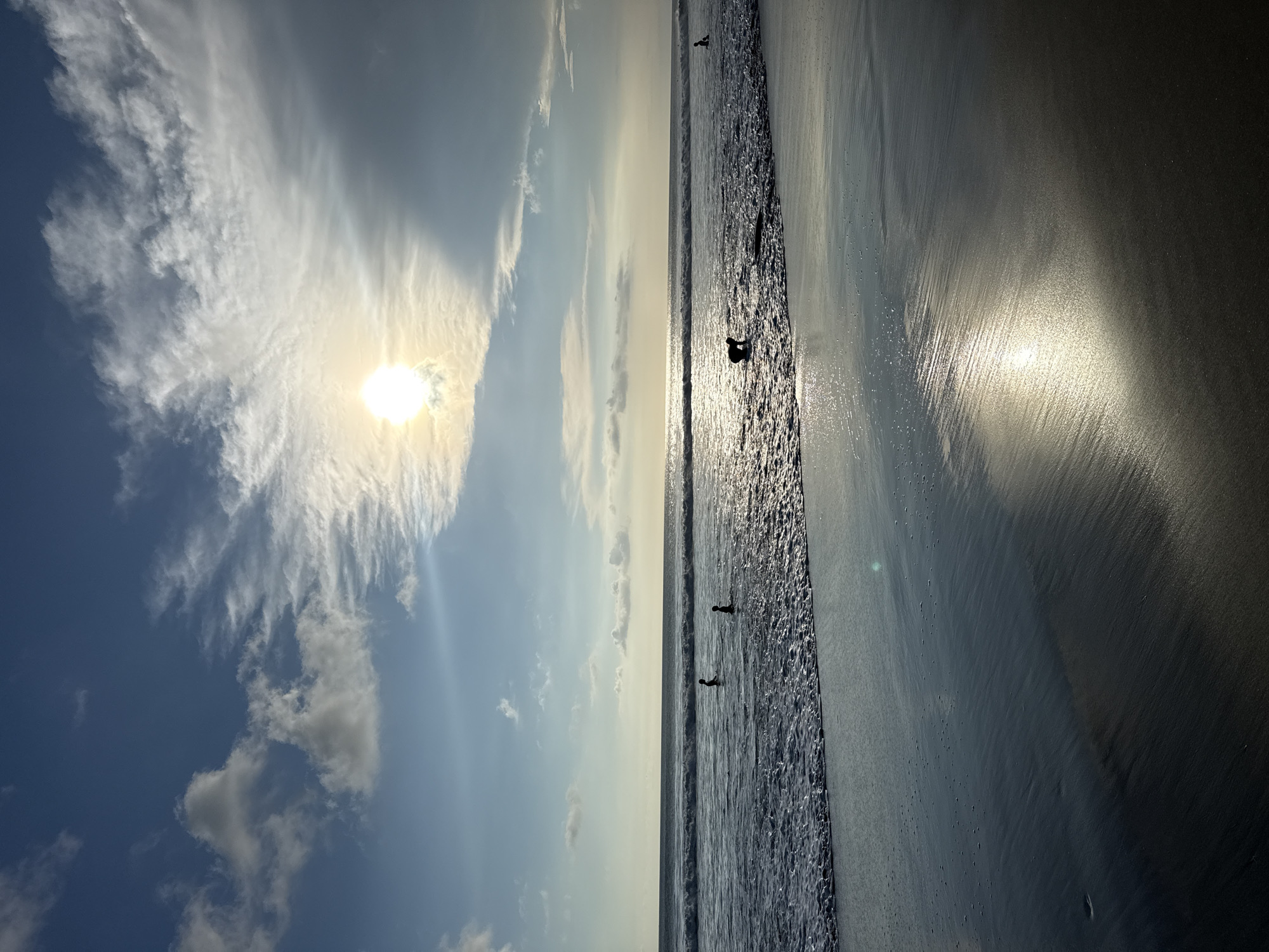 The image depicts a beach scene in Kuta, Indonesia. The sky is partially cloudy, with the sun visible through a thin cloud cover. The beach slopes towards the ocean, with the wet sand reflecting sunlight. Several indistinct figures are present in the water near the shoreline. The image orientation is vertical.