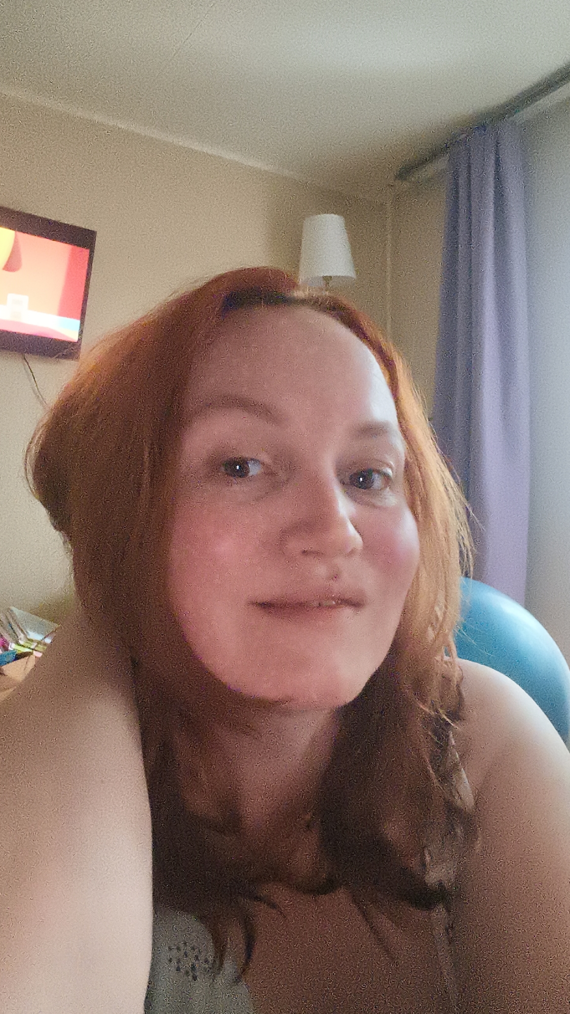 A woman with red hair poses for a selfie indoors. In the background, a television is mounted on a wall next to a table lamp, a blue curtain, and a blue exercise ball. The location is Unknown, Unknown.