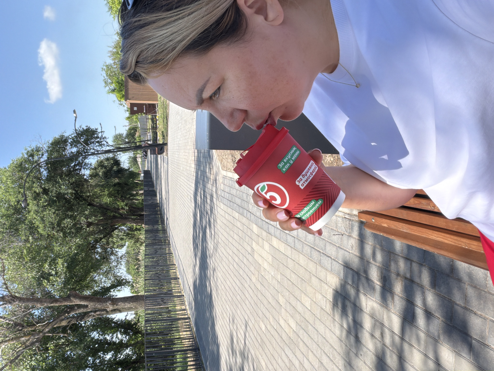 The media file shows a person in a close-up shot, holding a red paper cup and drinking from it. The person has blonde hair and is wearing a white t-shirt and red shorts. The cup has some text in Russian, including "За вкусным, как у мамы" and "За душевной атмосферой". A small green logo is also visible on the cup. The background appears to be an outdoor setting with a paved walkway, trees, and a blue sky with a few clouds, suggesting it is daytime and pleasant weather. The scene is set in Leningradskaya, Russia. The person's face is angled downwards, focused on drinking from the cup, with a serene expression. The lighting indicates it is a sunny day, casting shadows on the ground. The overall mood seems relaxed and casual.