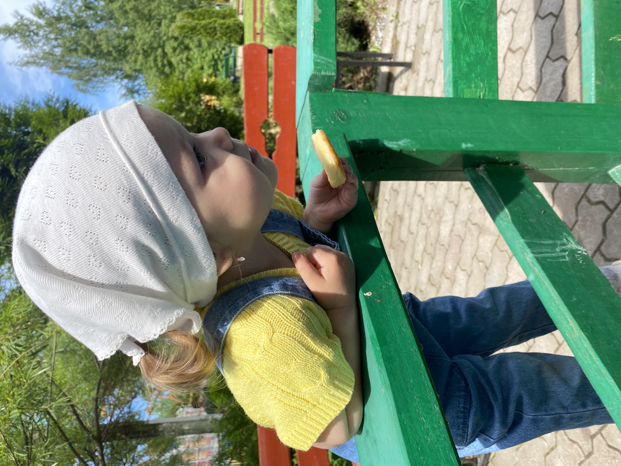 A young child is captured outdoors, seated or leaning on a robust green wooden structure, possibly playground equipment or a bench. The image is framed from a slightly elevated perspective, looking up towards the child's profile. The child wears a white, intricately patterned knitted or lacy head covering, a bright yellow cable-knit sweater, and blue denim overalls. Light-colored hair is visible at the back of the head, beneath the covering. The child's blue eyes are visible, looking upwards and to the left. In the right hand, the child holds a light-colored, rectangular piece of food, resembling an apple slice or a piece of cheese.
The setting is an outdoor, green environment. Abundant green foliage, including trees and bushes, fills the background. A distant red and green striped wooden structure is also discernible. The ground beneath the main green structure is paved with light and dark grey interlocking bricks laid in a wavy pattern. The scene is brightly lit by natural sunlight, casting clear shadows, indicating a sunny daytime. The child's expression appears calm and contemplative, focused upwards. No visible text is present in the image. The location context provided for this scene is Chaikovsky, Russia.