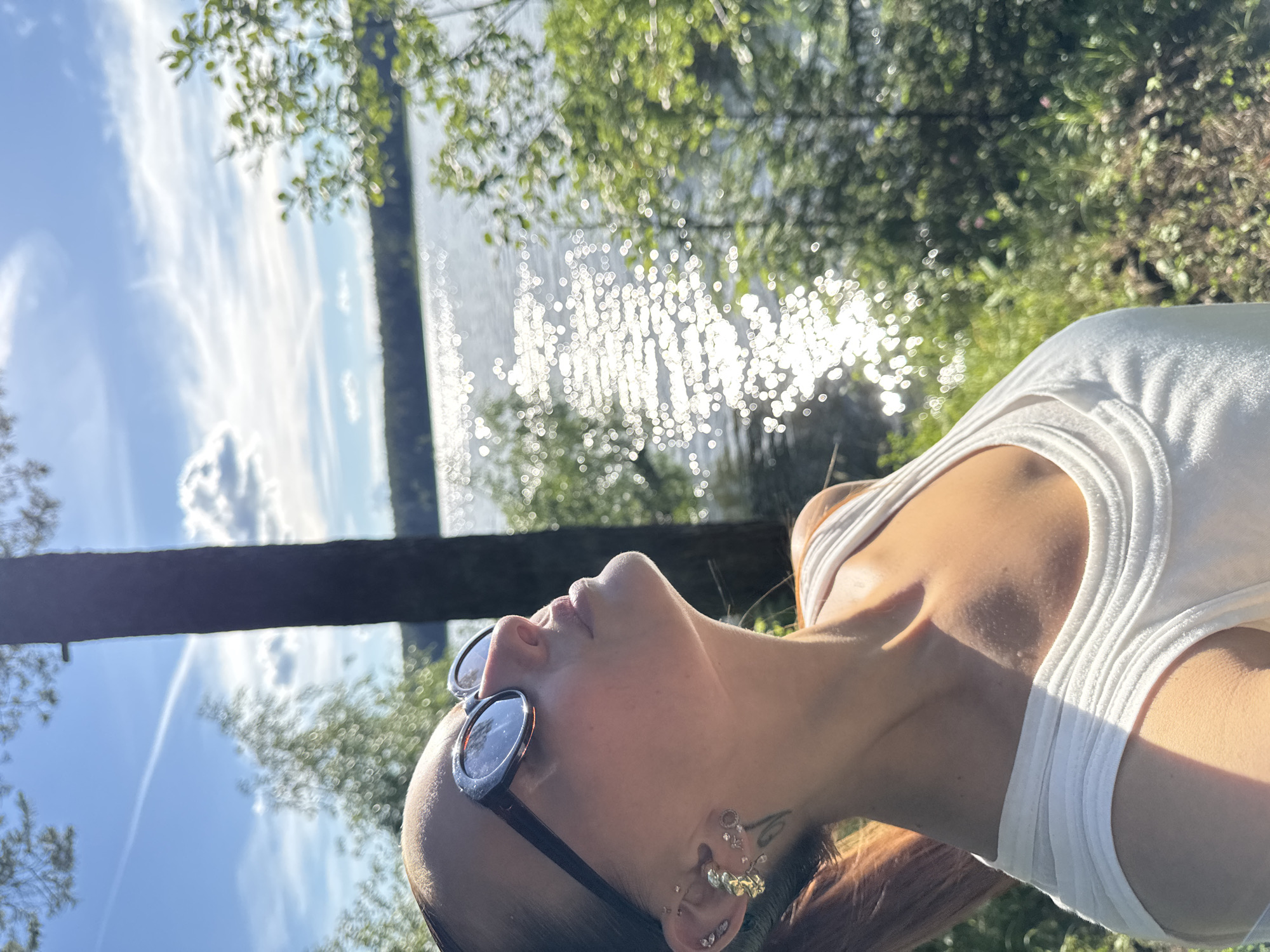 An outdoor, medium shot captures a woman in profile, gazing upward towards the sky. She is wearing sunglasses, a white tank top, and several earrings in her left ear. Her auburn hair is pulled back into a ponytail. Sunlight illuminates her face and décolletage. Behind her, a tree trunk stands prominently, partially obscuring a body of water that sparkles with reflected sunlight. The surrounding area is lush with green foliage. The location is Votkinsk, Russia.