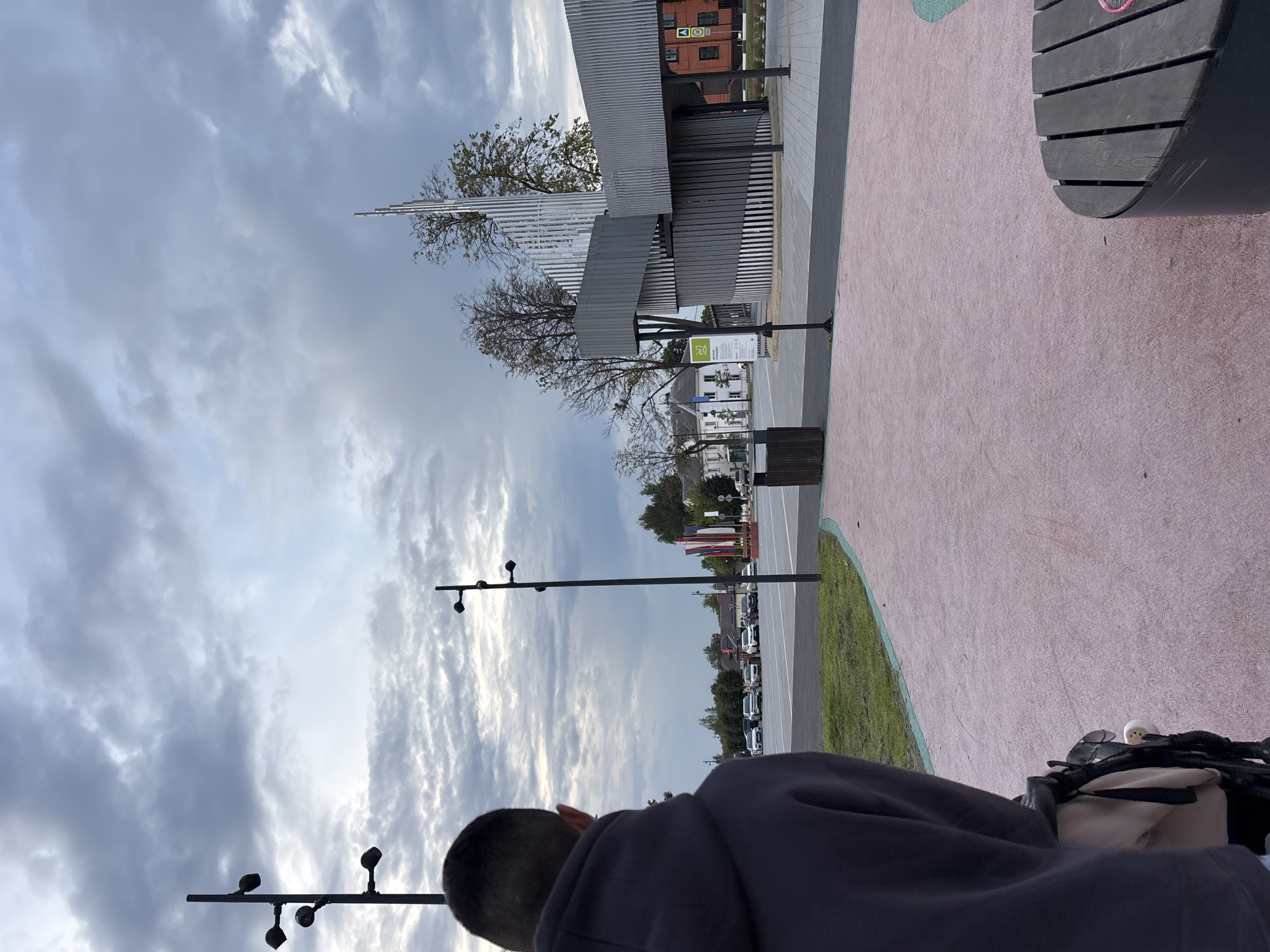 The image shows a view of the city of Klin, Russia, with a person in the foreground. The person is facing away from the camera, wearing a dark hooded sweatshirt. In the background, there is a modern architectural structure with a spiral design, possibly a pavilion or a monument. Several trees are visible, some with sparse foliage, suggesting it might be autumn. A parking lot with cars is also present. Flags are displayed in the distance. The sky is overcast with dramatic clouds. The ground in the foreground is a pinkish-colored surface, possibly a walking path or plaza, with a green border. A dark wooden bench is partially visible in the lower right corner. The overall scene appears to be a public space in the city.