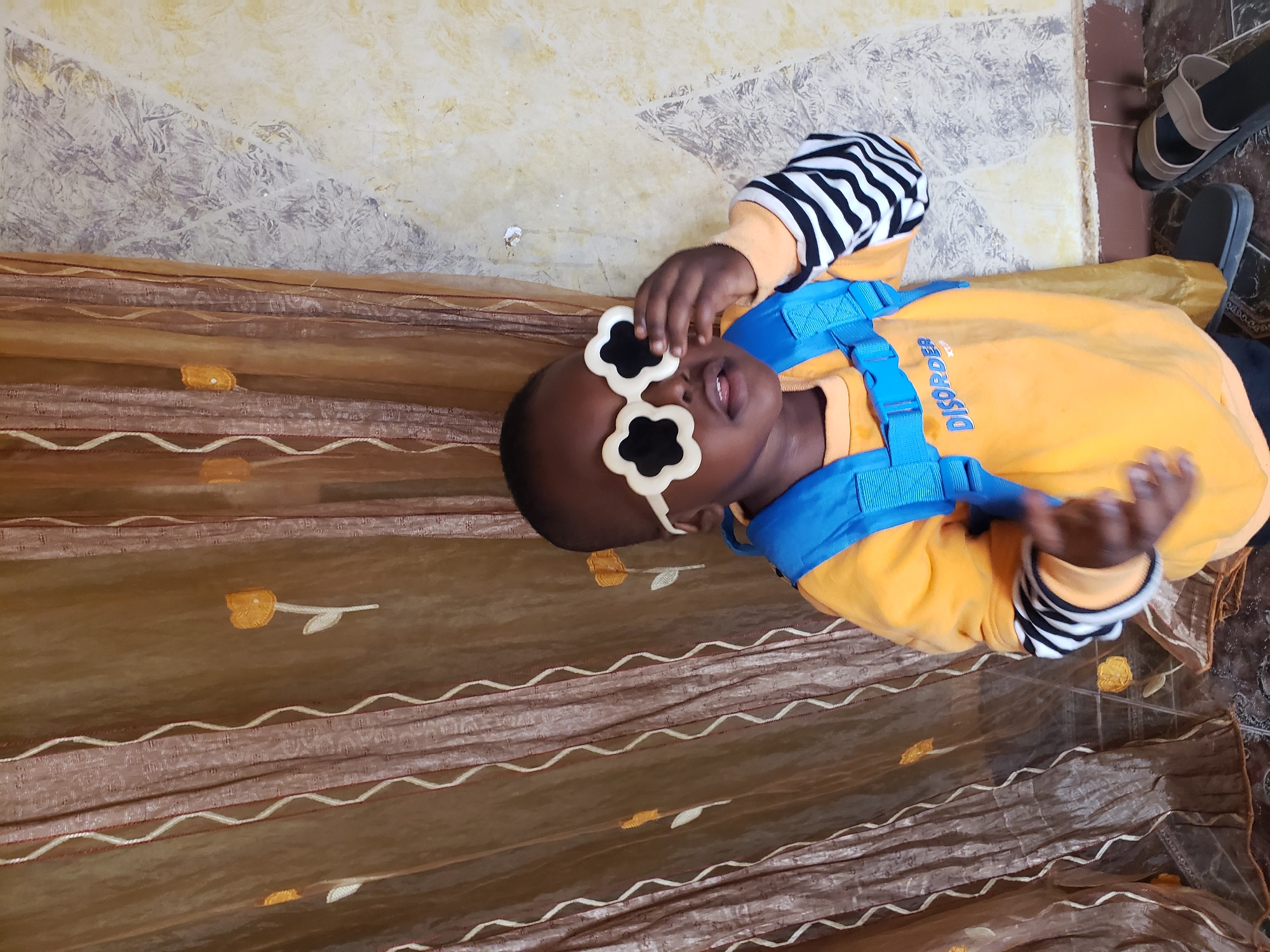 A young child, likely a boy, stands in front of a patterned brown curtain and a textured wall. The child is wearing a bright yellow long-sleeved shirt with black and white striped cuffs, a blue backpack, and distinctive white flower-shaped sunglasses. The child's right hand is raised, touching the sunglasses. The background curtain has an embroidered floral pattern in orange. The wall appears to have a distressed or textured paint finish in shades of cream and gray. Two pairs of sandals are visible on the tiled floor in the foreground. The scene appears to be indoors, possibly in a home in Sabon Tasha, Nigeria.