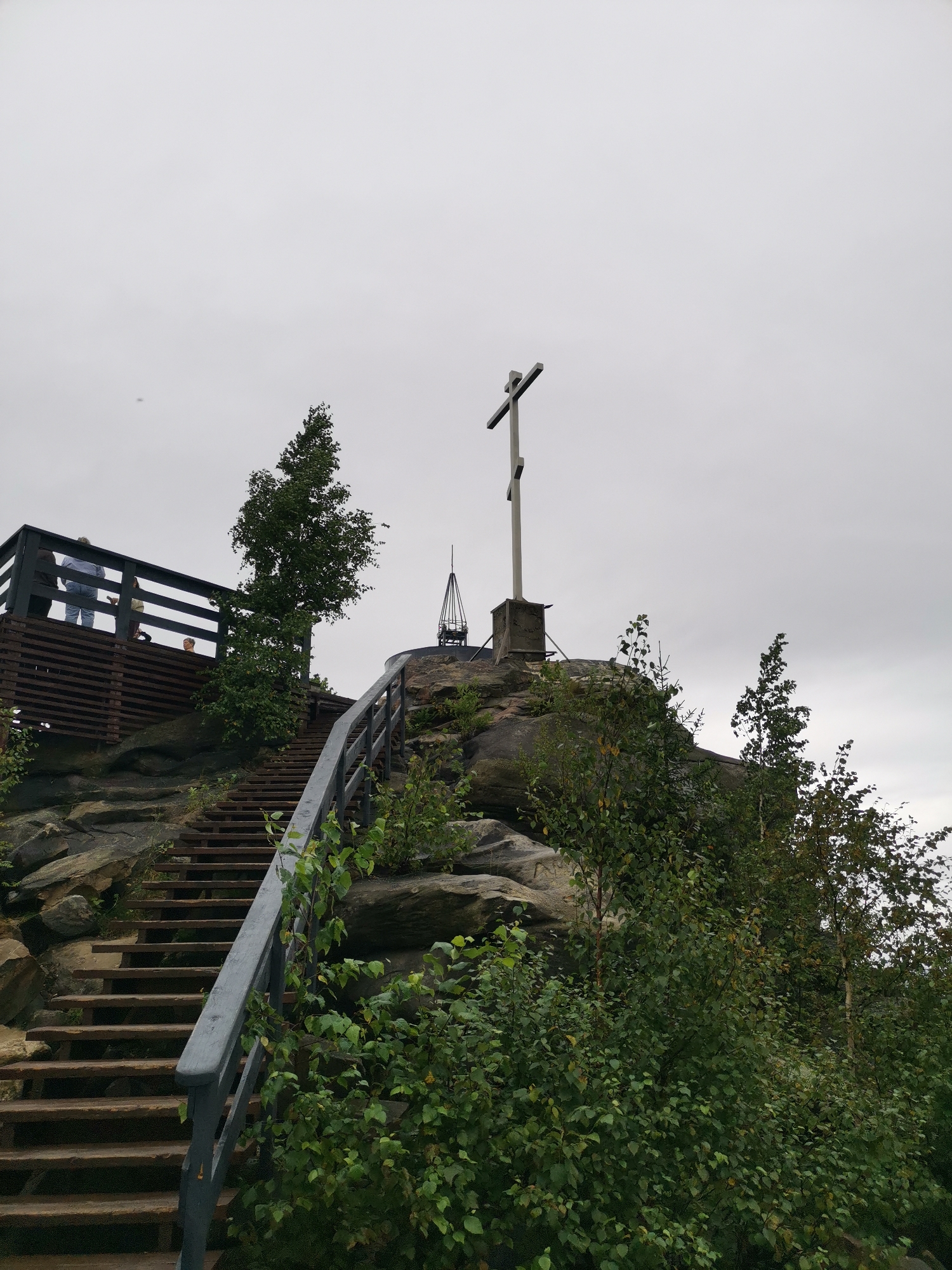This image captures a view from a wooden staircase leading up to a rocky outcrop with a large metal cross. The cross is situated on a stone base, and behind it, a slender, dark metallic spire can be seen. To the left, a wooden viewing platform with dark railing is visible, with several individuals standing on it, some looking out at the surroundings. The platform has a slatted wooden fence. Lush green foliage, including trees and bushes, grows around the rocks and along the staircase. The sky is overcast and grey. The scene is located at Mount Kizelovsky in Gubakha, Russia, a popular tourist destination known for its natural beauty and historical significance.