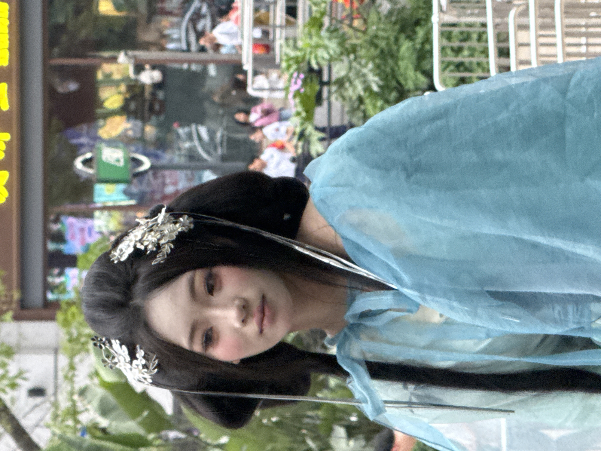 A portrait captures a woman in traditional Chinese attire, specifically hanfu, positioned in Yuzhong District, China, as discerned from the backdrop. The woman's hair is styled in a traditional bun adorned with silver-colored hairpins. She wears a flowing, light blue gown. Her makeup includes rosy blush.
In the background, pedestrians are visible behind a glass storefront, along with greenery and a metal railing. The image appears to be taken outdoors during daylight. A yellow sign is partially visible in the upper left corner.