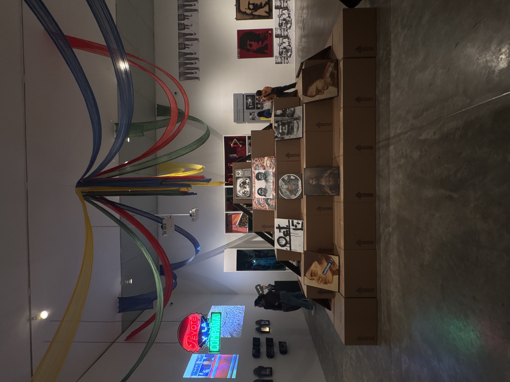 This is a photograph of an art exhibition. In the foreground, a tiered display constructed from cardboard boxes holds various framed artworks. The boxes are stacked in a pyramid shape, with the artworks placed on each level. One prominent artwork on the lower tier features a portrait of a person with "LISTERINE" written on a blue strip across their face. Above this, other framed pieces are visible, including a square artwork with "Oct 15" written on it and religious iconography.
Further back, a neon sign in red and green reads "Peyote y la Compania". To the left of the sign, a large screen displays an image, possibly a video. On the walls in the background, several other artworks are displayed, including portraits and what appear to be prints of legs.
Several people are present in the scene. Two individuals are standing in the mid-ground, looking at artworks. Another person is standing on the right side of the display, looking at a piece. The ceiling is adorned with draped colorful fabric in yellow, green, red, and blue. The environment appears to be an indoor exhibition space with white walls and a concrete floor. Some areas of the floor reflect the colored lights from the fabric.