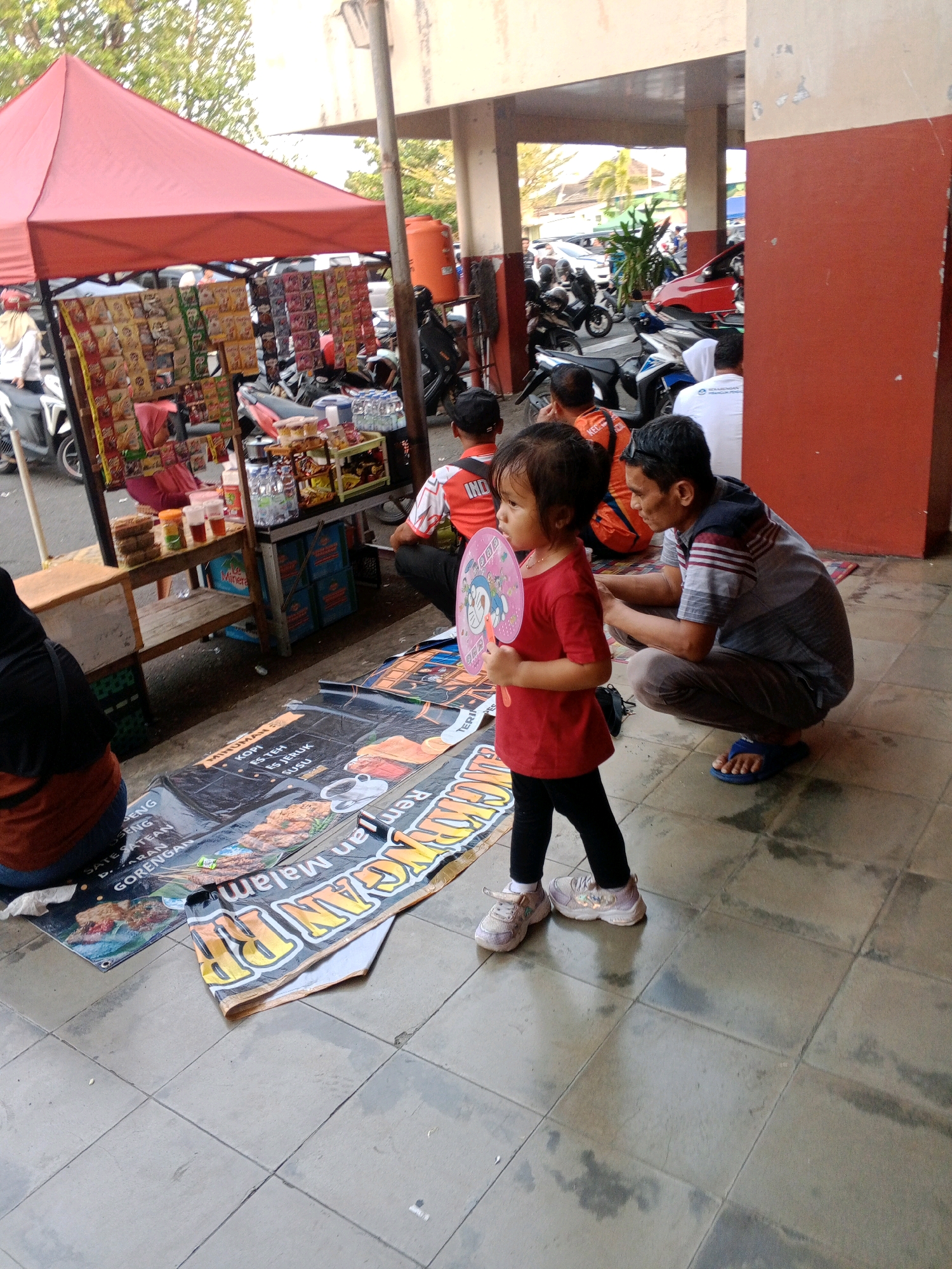 In Wuryorejo, Indonesia, a young girl in a red shirt and black leggings stands on a tiled outdoor floor, holding a pink fan with a cartoon character. She is facing left, looking away from the camera. To her right, a man in a grey and striped shirt and brown pants squats down, looking at his phone. Behind them, a red awning covers a stall displaying various packaged food items. Motorbikes are parked in the background, along with other people in casual attire. Several large banners are spread on the ground in front of the stall, displaying text and images, one of which appears to be an advertisement for food. The environment suggests a street-side market or food stall area.