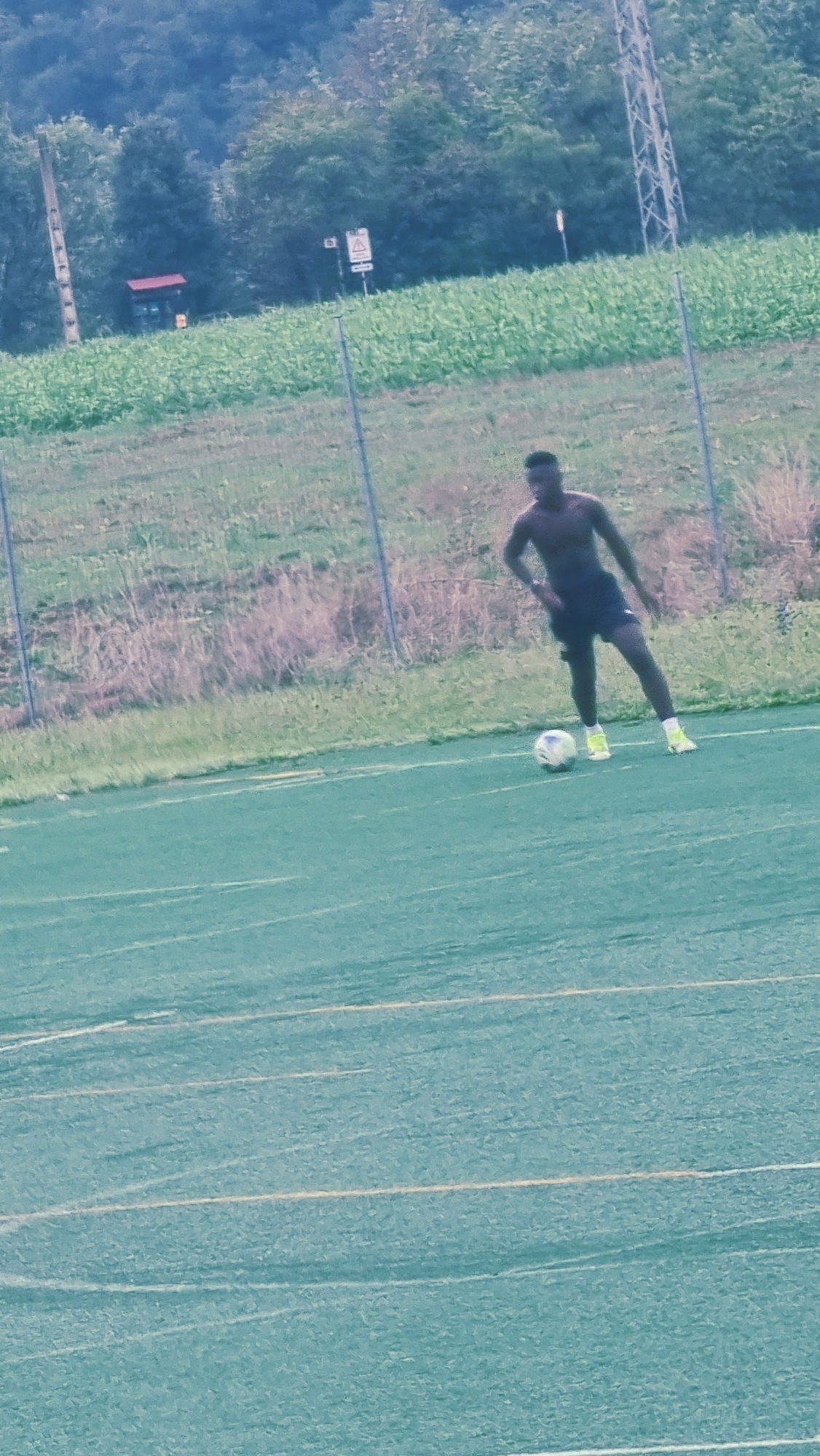 A shirtless Black male is playing soccer on a green artificial turf field. He is wearing dark shorts and yellow cleats. The soccer ball is near his left foot. In the background, there is a chain-link fence, dry grass, and a field of green crops. Beyond the field, there are trees and a power line tower. The scene appears to be in Sant Joan les Fonts, Spain, suggested by the surrounding natural landscape and infrastructure.
