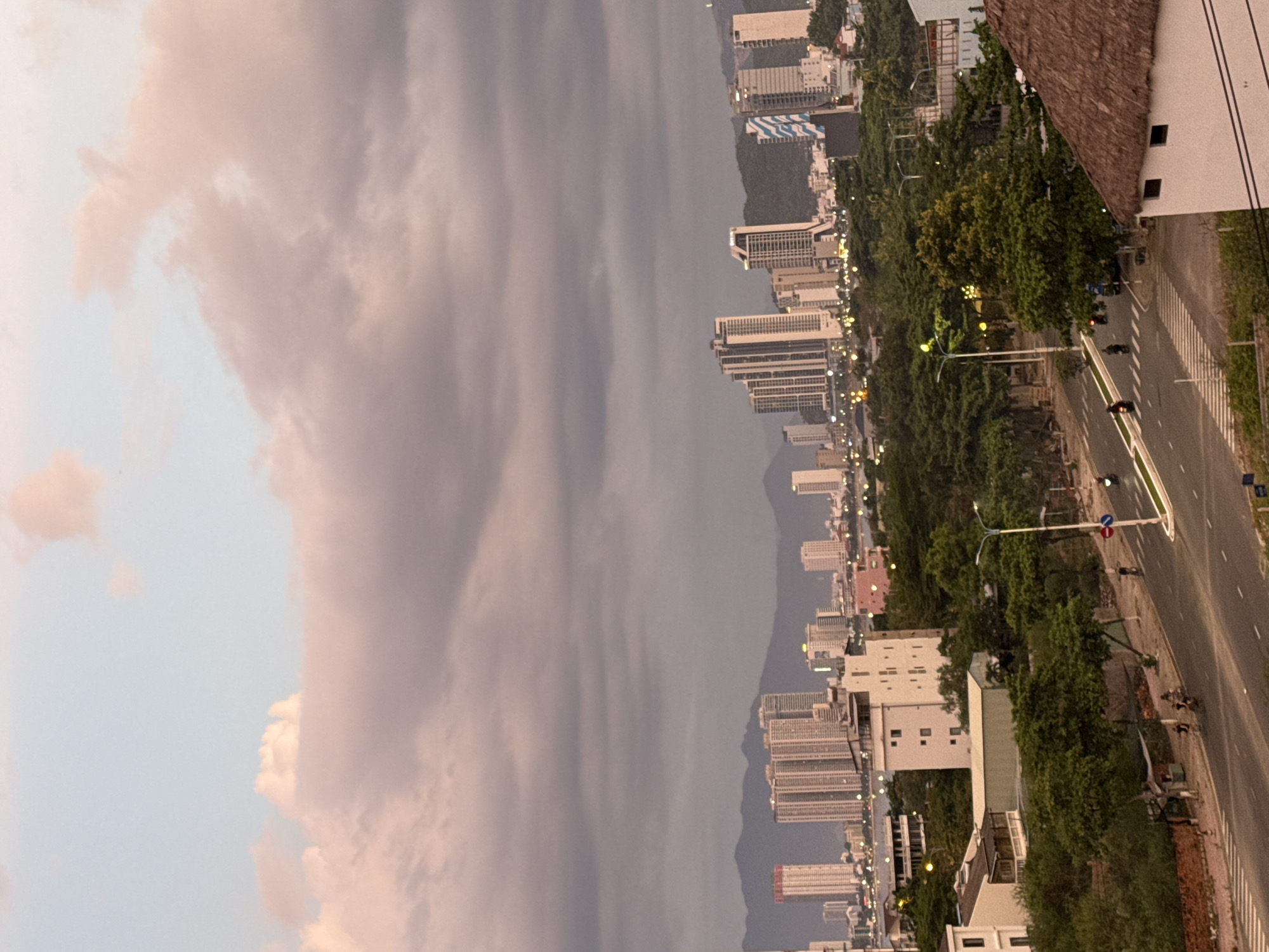 The image depicts a coastal city, likely Nha Trang, Vietnam, as dusk settles. The sky is filled with dramatic, pink and gray clouds, suggesting an impending storm or the aftermath of one. Dominating the scene is a dense cluster of tall, modern buildings that line the coast, their windows reflecting the dim light. In the foreground, a road runs alongside the buildings, with a few motorcycles visible, indicating ongoing activity despite the subdued lighting. Trees are interspersed throughout the cityscape, adding a touch of greenery to the urban environment. The overall mood is one of transition, with the fading light and heavy clouds creating a slightly dramatic or melancholic atmosphere. The architecture suggests a developed tourist area, with the presence of the road and vehicles implying a functional urban infrastructure. No discernible text is visible in the image.