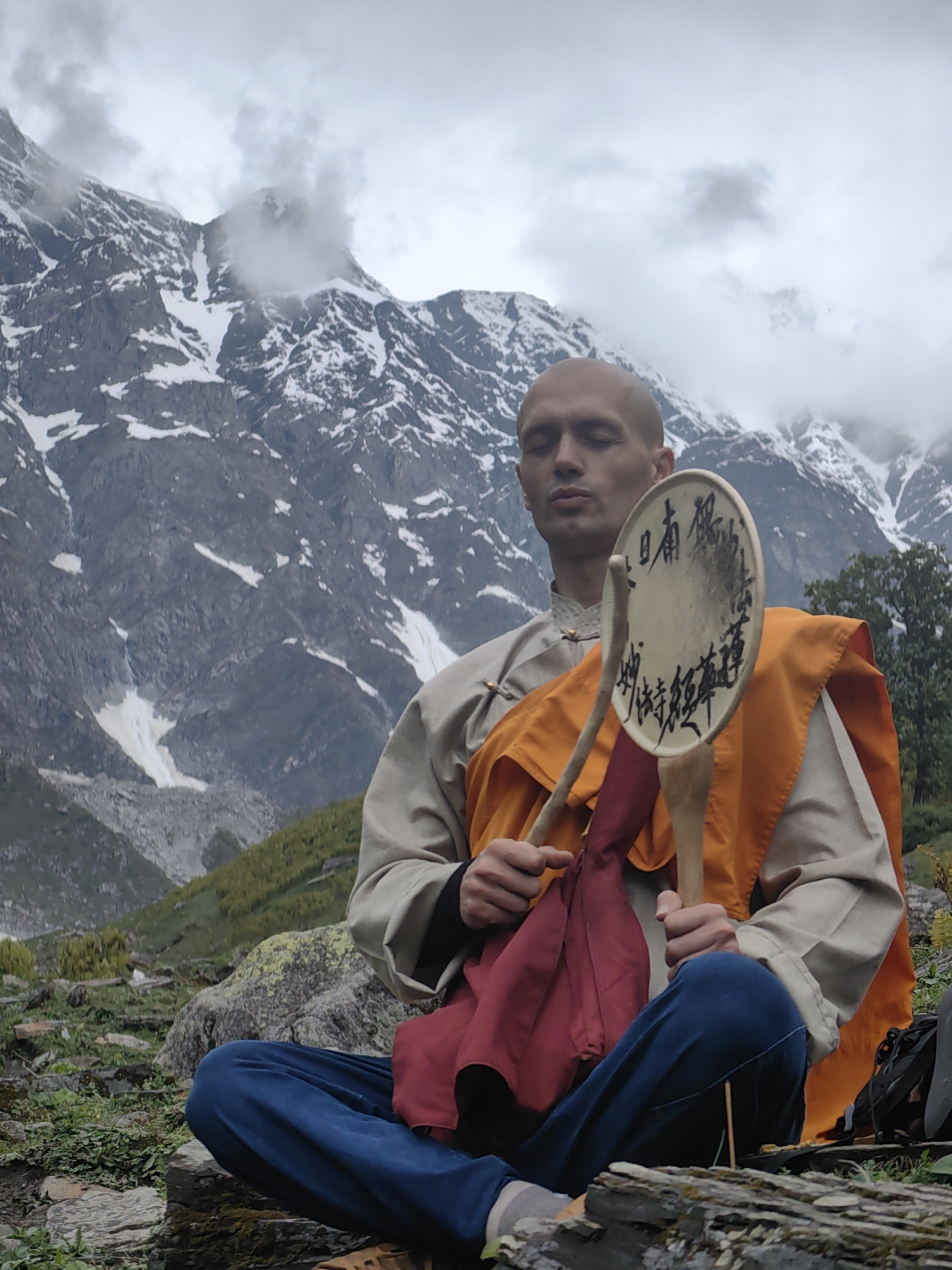 A man, likely a Buddhist monk given his shaved head and saffron and tan robes, sits in meditation on a rocky, grassy slope. He is dressed in a tan robe with a dark undershirt, and an orange sash is draped over his shoulders. A burgundy cloth is tucked into his sash. He holds a round, wooden object, possibly a drum or frame, with two handles, on which some dark text, likely calligraphy, is visible. His eyes are closed in concentration.
Behind him, a dramatic mountain landscape unfolds under a cloudy, overcast sky. The mountains are rugged and imposing, with snow clinging to their upper reaches. Patches of green grass and shrubs cover the lower slopes, interspersed with large rocks. A few trees are visible in the middle ground to the right.
The weather appears cool and possibly damp, with mist or low-lying clouds partially obscuring the mountain peaks, suggesting a time of day when the light is diffused. The overall atmosphere is serene and contemplative, evoking a sense of solitude and spiritual practice within a majestic natural setting.
The visible text on the wooden object appears to be in Asian script, possibly Chinese or Tibetan, written vertically.