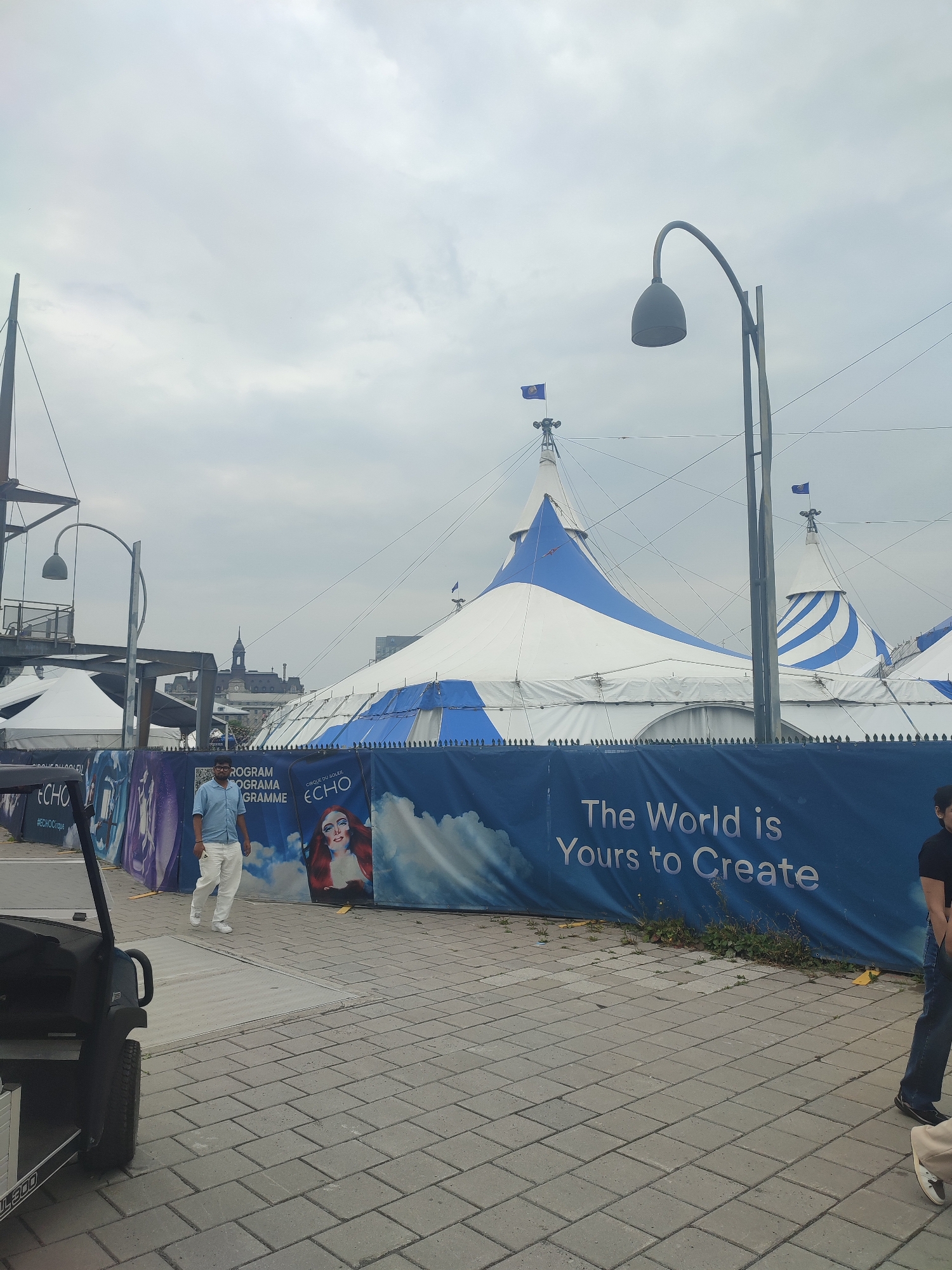 This is a medium shot, eye-level, outdoor, daytime, slightly overcast photograph of a Cirque du Soleil "Echo" tent setup in Montreal, Canada. The main subject is a large, white and blue striped circus tent with multiple peaked sections. In the foreground, a dark-colored golf cart is parked on the left side of the frame, partially obscuring a section of the paving stones. A man wearing a light blue short-sleeved shirt and white pants is walking on the paving stones towards the right of the frame. Behind him, a blue banner runs along a fence, featuring the Cirque du Soleil logo and the text "The World is Yours to Create." Several other smaller white tents are visible behind the main tent and to the left. A streetlamp with a curved arm and a conical shade stands to the right of the frame. In the background, the Montreal skyline is visible, including a building with a distinctive tower. The overall scene suggests an outdoor event or performance space.