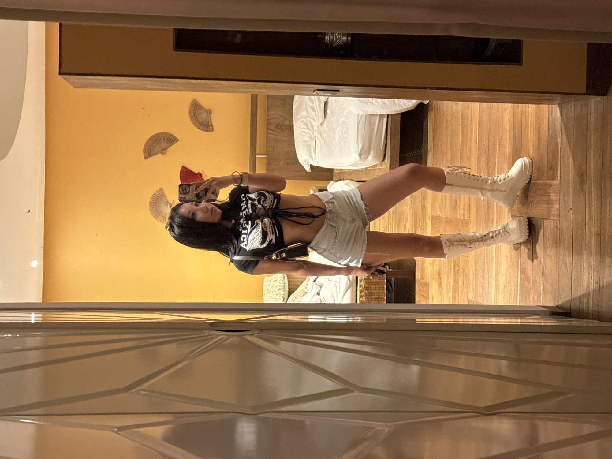 A woman is taking a mirror selfie in what appears to be a hotel room. She is wearing a black crop top with a lace-up front and a graphic print, a pair of light-wash denim shorts, and tall white lace-up boots. She has long, dark hair and is holding a phone to take the photo. The room has a warm, orange-toned wall with decorative fans mounted on it. A bed with white linens is visible in the background, along with wooden furniture and a wooden floor. A portion of a mirrored closet door is also in the frame. The location is identified as North Kuta, Indonesia.
