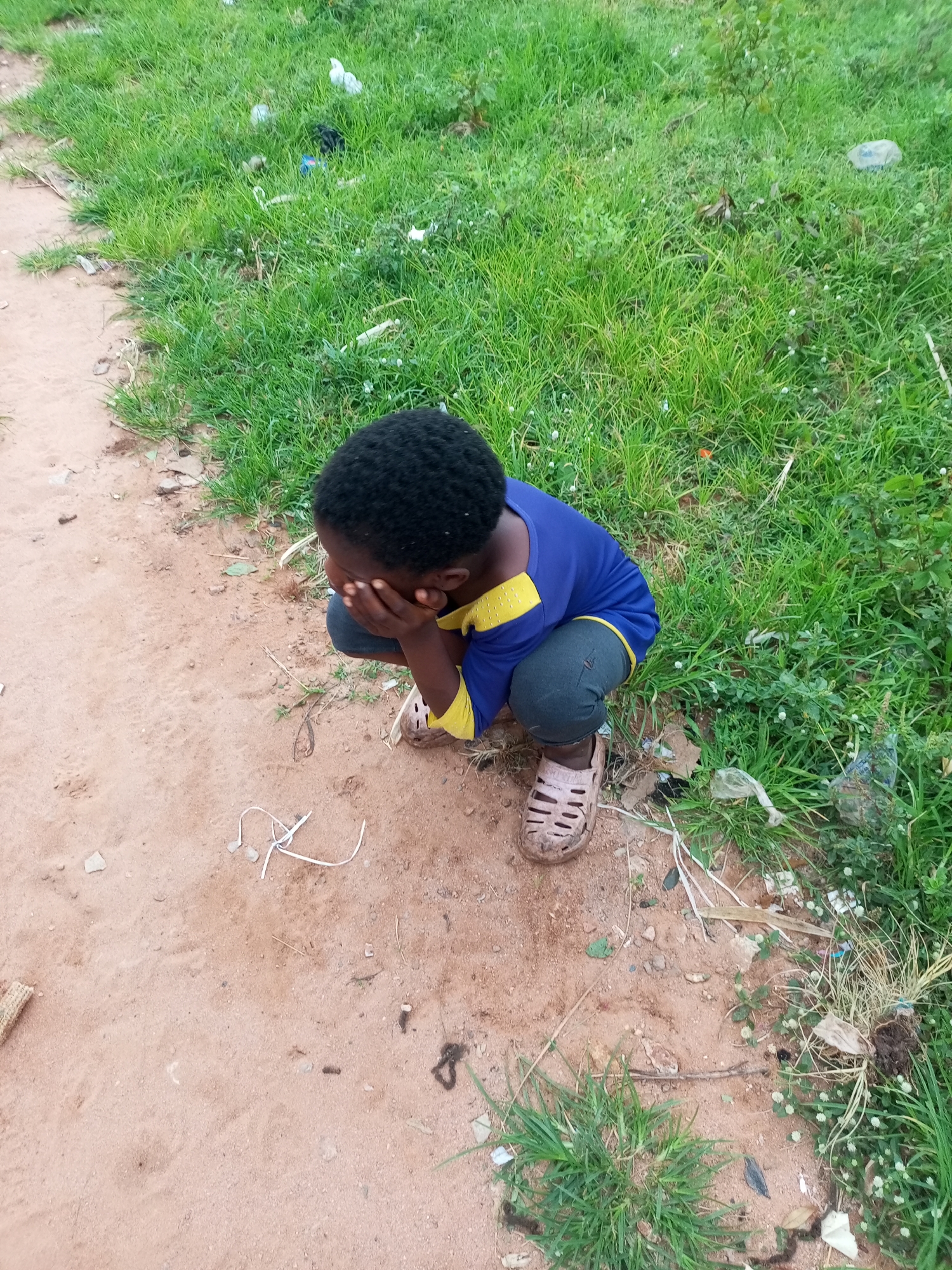 A young Black child is crouching on sandy ground next to a patch of green grass in what appears to be an outdoor, possibly urban, environment in Tempure, Nigeria. The child is wearing a blue shirt with yellow accents on the collar and sleeves, and dark pants. They have their hands covering their face, suggesting they might be crying or hiding. On their feet, they are wearing light-colored, open-toed sandals. Scattered on the ground around the child are small pieces of debris, including white plastic-like material, a few pebbles, and some dry grass. The grass area to the right of the child appears somewhat unkempt, with a few visible pieces of litter interspersed within the foliage.