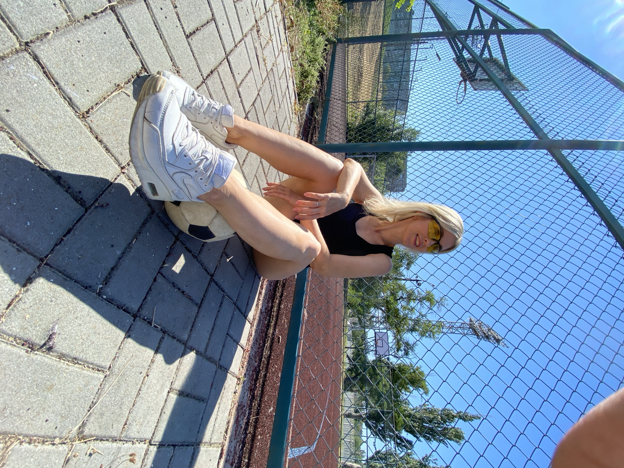 A blonde woman wearing sunglasses and a black tank top sits on a paved area next to a chain-link fence. She has her legs extended and a soccer ball is resting under her right leg. She is wearing white sneakers. In the background, there is a basketball hoop and a sports field. The sky is blue and clear.