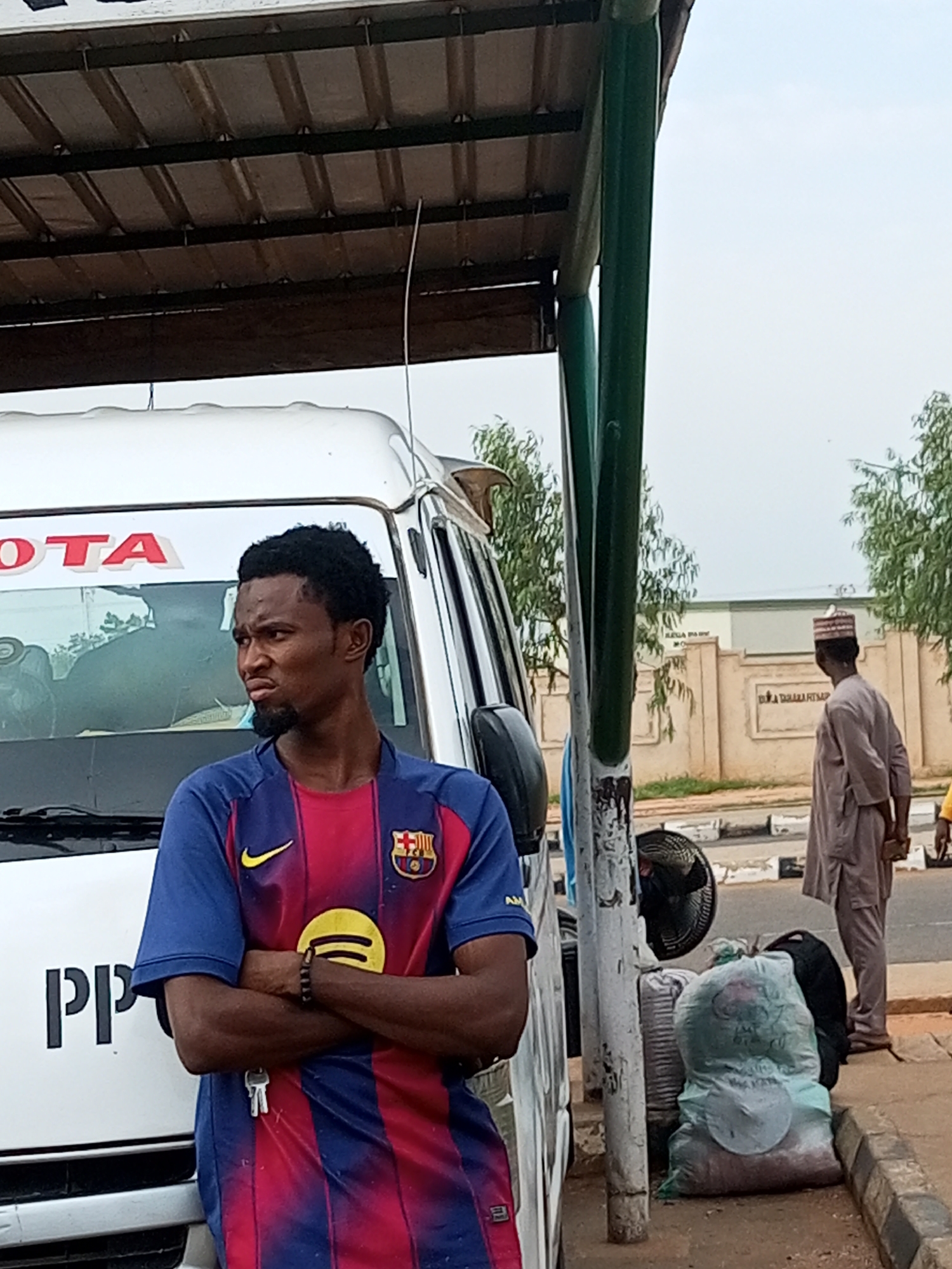 A young man stands with his arms crossed in front of a white Toyota van, wearing a blue and red striped shirt with a Barcelona FC crest. He has a short beard and dark, curly hair. To the right, an older man in a light-colored kaftan and a patterned cap stands looking away from the camera. Several large bags and a black fan are placed on the ground near a metal pole. The background shows a beige wall with text that is difficult to read, trees, and a cloudy sky, suggesting an outdoor setting in Gombe, Nigeria. The scene appears to be at a bus stop or a commercial area.