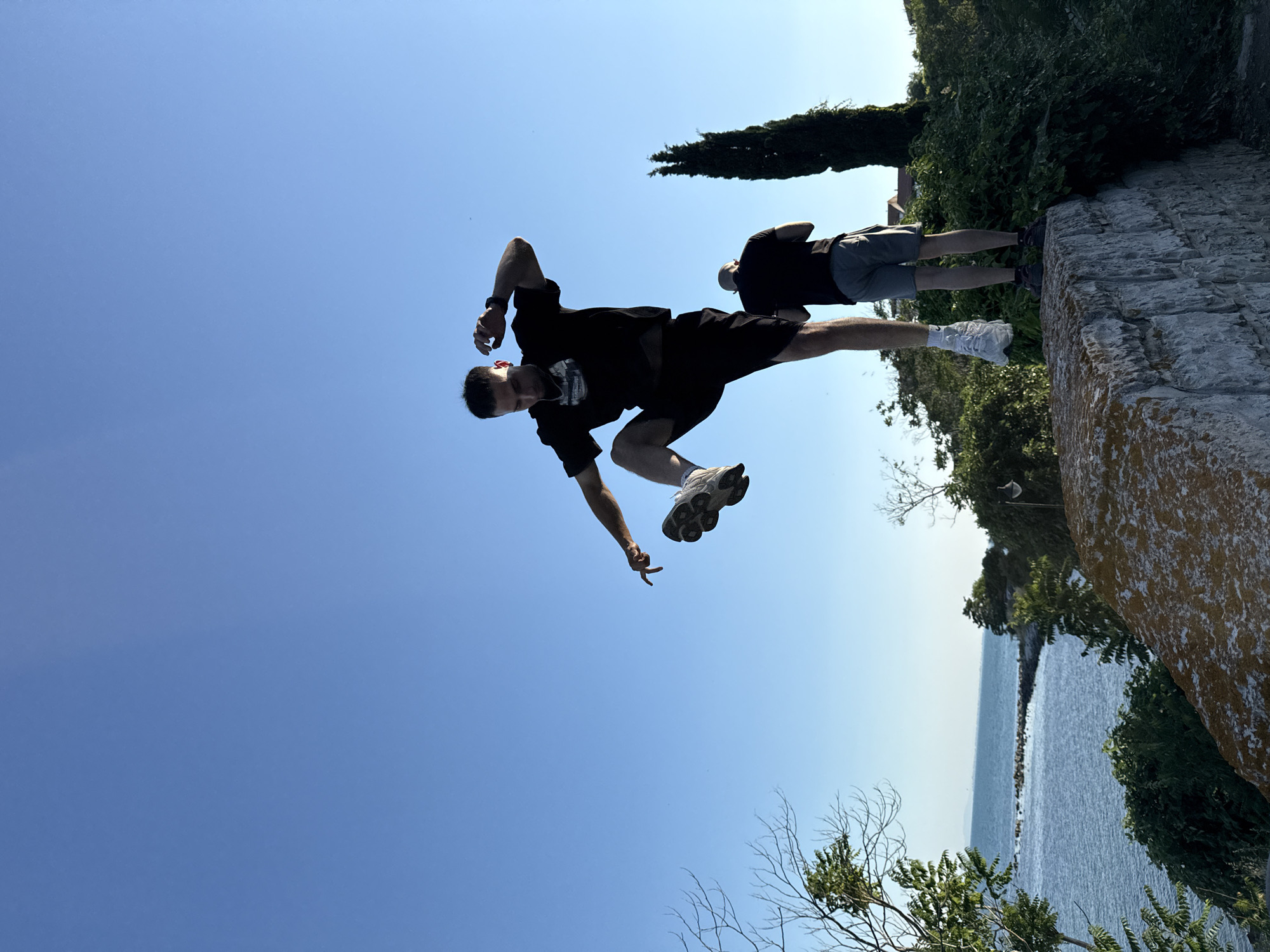 This media file features two individuals engaged in an activity against a backdrop of a clear blue sky and a body of water. One person is captured mid-air, seemingly in the act of jumping or leaping. This individual is wearing a black t-shirt, black shorts, and white athletic shoes. Their body is oriented horizontally, with one arm extended and the other bent at the elbow. The other person is standing further back and to the right, facing away from the camera and appearing to be observing or waiting. They are dressed in a dark t-shirt and grey shorts.
The scene is set outdoors, with lush green foliage and trees visible along the top edge of the frame, suggesting a natural or park-like environment. A textured, rock-like surface occupies the right side of the image, adding to the sense of a natural landscape. The sea is visible in the distance, with a clear horizon and calm waters. The time of day appears to be sunny, indicated by the bright sky and the lighting on the subjects. The overall mood is energetic and playful, conveyed by the dynamic pose of the jumping individual. There is no visible text in the image. The setting is consistent with the coastal environment of Nesebar, Bulgaria.