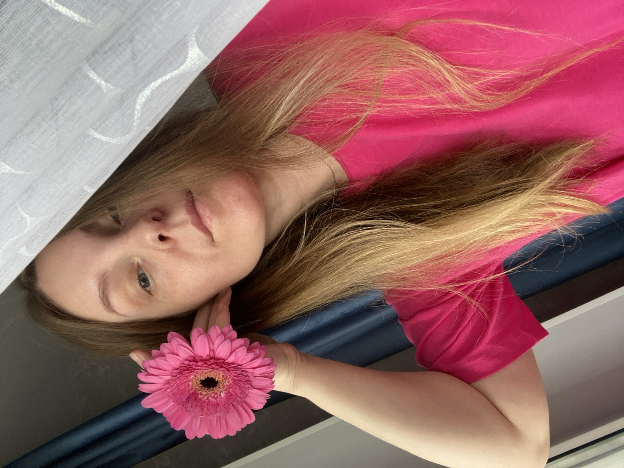 A woman with light skin, light brown hair, and blue eyes is holding a pink gerbera daisy near her face. She is wearing a bright pink t-shirt. Her hair is long and partially covers her face. The background consists of a white curtain on the left side, a dark blue surface behind her neck and shoulder, and a white surface below her arm. The photo appears to be taken indoors, possibly near a window. The location of the photo is not discernible from the image and cannot be identified as Noviy, Russia.