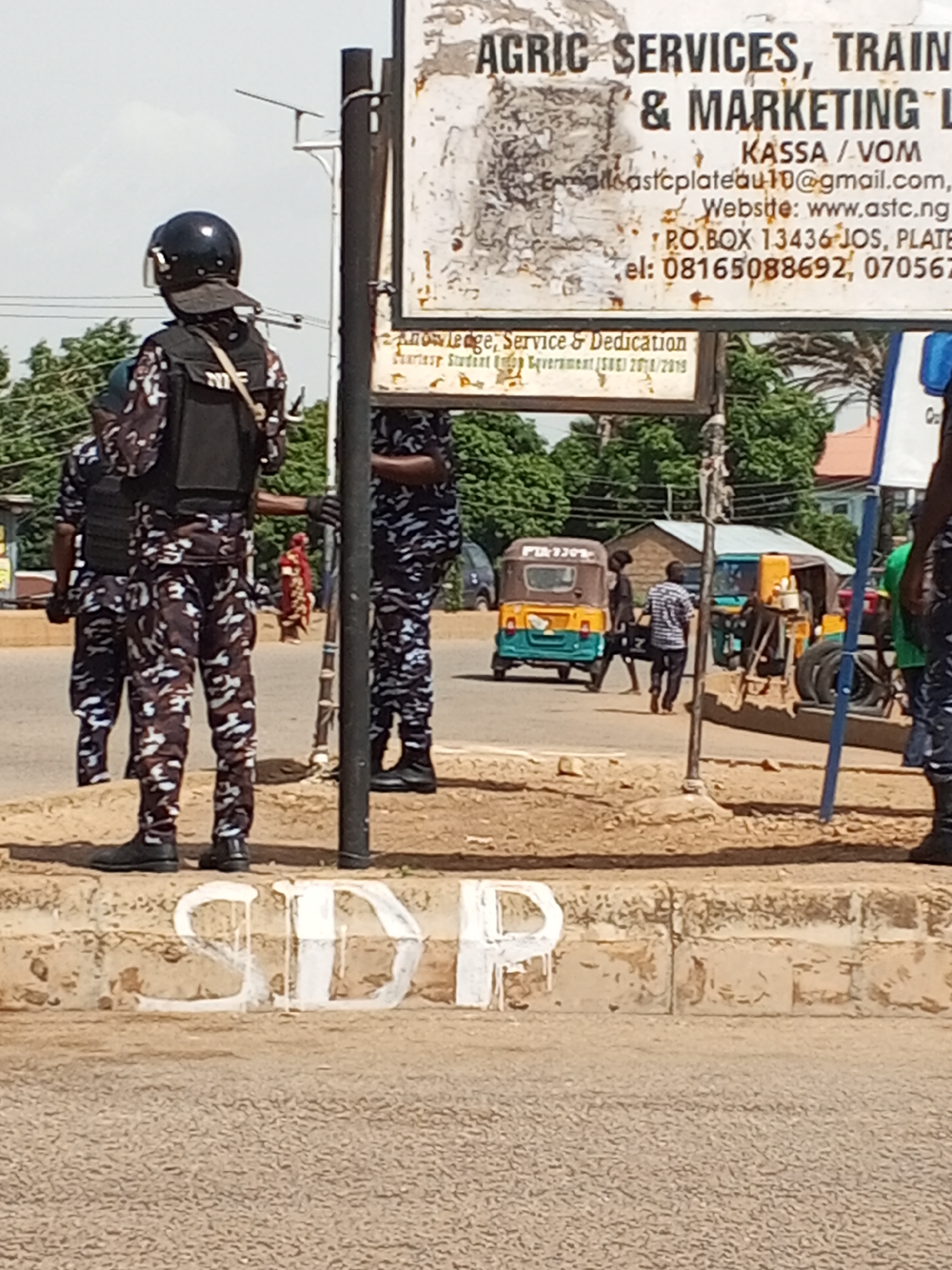 The image depicts a street scene in Bukuru, Nigeria, featuring several individuals in what appears to be security or law enforcement attire. In the foreground, a person wearing a camouflage uniform and a black helmet with a visor stands facing away from the camera. Their uniform consists of a patterned shirt and pants, and they are also wearing a black vest. To their left, another individual in a similar uniform is partially visible, also facing away. To the right, another person in the same camouflage uniform is visible from the waist down, wearing black boots. In the background, there are various elements typical of a Nigerian street. A signpost with faded writing is prominent, listing "AGRIC SERVICES, TRAIN & MARKETING L," a Gmail address, a website, a P.O. Box, and phone numbers. Several auto-rickshaws, also known as Keke Napep, are present, with one in the middle of the frame that is blue and yellow. There are also stacks of tires and a person in a striped shirt walking in the background. The environment appears to be outdoors, with a paved road and a dirt or gravel-covered area in the foreground where the word "SDP" is painted in white letters on a concrete curb. Trees and buildings are visible in the distance. The overall impression is of a public space in an urban or semi-urban setting.