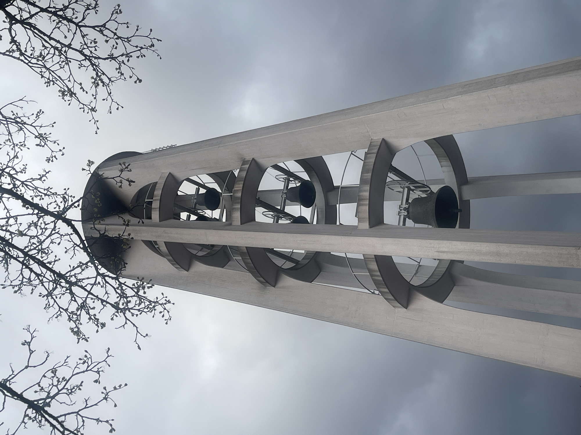 The image shows a view of the tower of the Evangelische Kirche Alt-Saarbrücken (Protestant Church of Old Saarbrücken) in Saarbrücken, Germany, from a low angle. The camera is looking upward at the bell tower structure, which is constructed of concrete and has an open design, revealing the bells inside. A tree branch with budding leaves is visible in the foreground on the left side of the frame, partially obscuring the tower. The sky is overcast and grey, suggesting a cloudy day. No people or interactions are visible in the image.