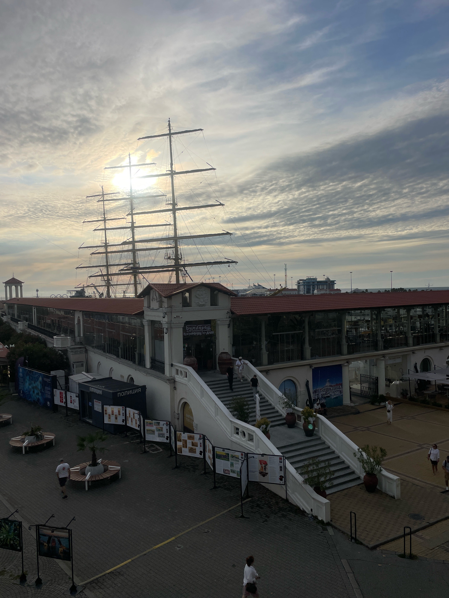 The image captures a view of the Sochi Sea Port in Russia, featuring a large sailing ship docked prominently. The sun is partially obscured by clouds, casting a soft light over the scene. In the foreground, a wide, white staircase leads up to a building with a red-tiled roof and large glass windows, suggesting it might be a restaurant or commercial establishment. The building is adorned with a sign that reads "БУМБАРАШ РЕСТОРАН ОТ ДУШИ" (BUMBARASH RESTAURANT FROM THE SOUL). Various informational displays are set up on stands along the walkway and near the entrance to the building.
To the left of the stairs, a pedestrian area is visible with circular benches surrounding small palm trees. A man in a white t-shirt and shorts is walking away from the camera in this area. Further to the left, a dark blue kiosk with the word "ПОЛИЦИЯ" (POLICE) is visible.
The sailing ship, with its multiple masts and rigging, dominates the upper portion of the image. In the background, other buildings and structures of the port can be seen, along with the sea and a lighthouse in the distance. Several people are scattered throughout the scene, walking or standing, indicating a moderate level of activity. The sky is filled with textured clouds, typical of a partly cloudy day. The overall atmosphere suggests a pleasant daytime scene at a tourist-oriented location.