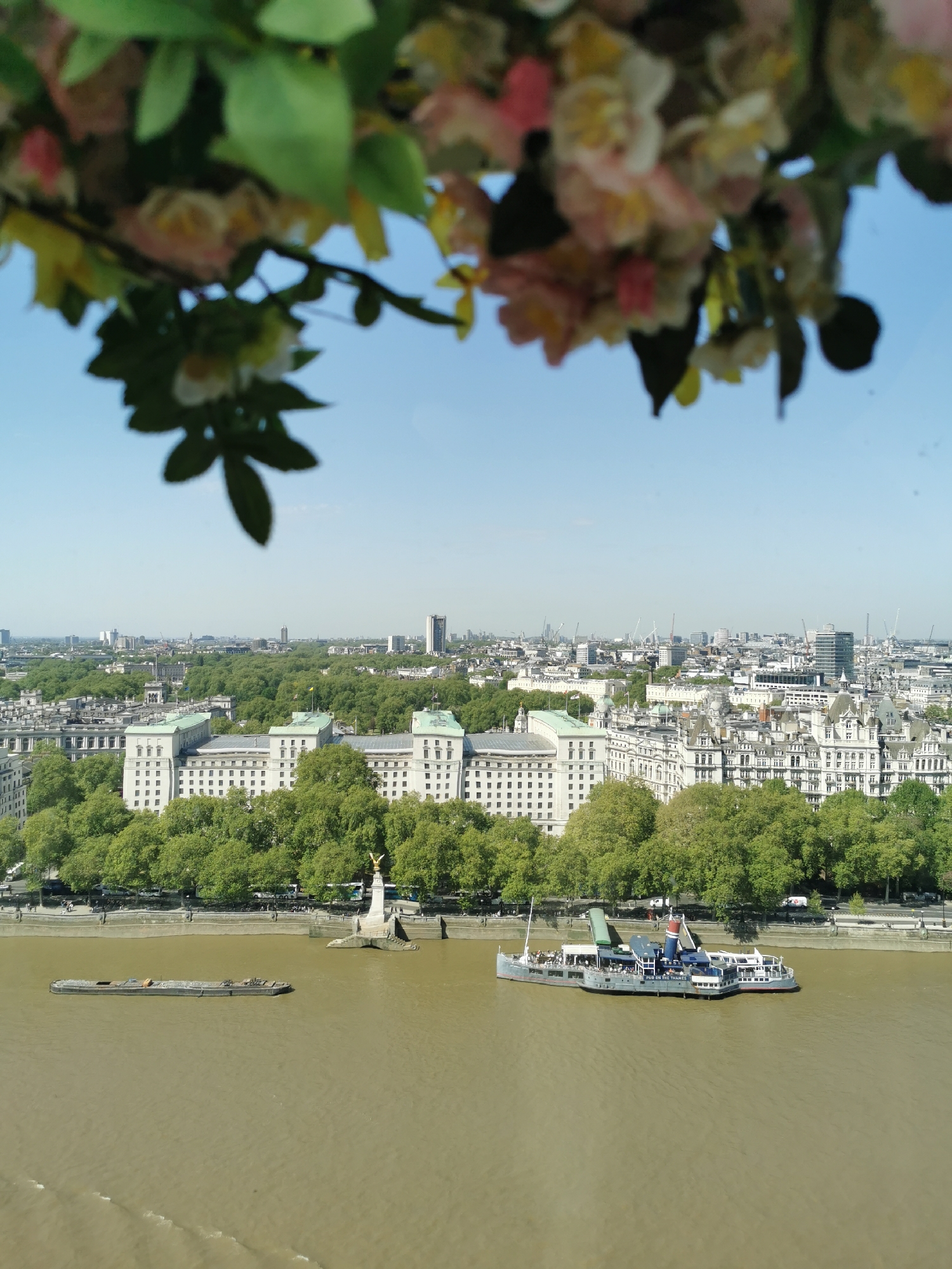 The image depicts a view of London, likely from a high vantage point such as the London Eye. The River Thames is visible with two boats, one a barge and the other a passenger boat with the words "PUB ON THE THAMES". A statue of a figure with outstretched wings is visible on the river bank. Behind the river, a line of trees is adjacent to white buildings. In the background, the London skyline is visible under a clear, light blue sky. In the foreground, blurred pink and white flowers and green leaves frame the top of the shot.