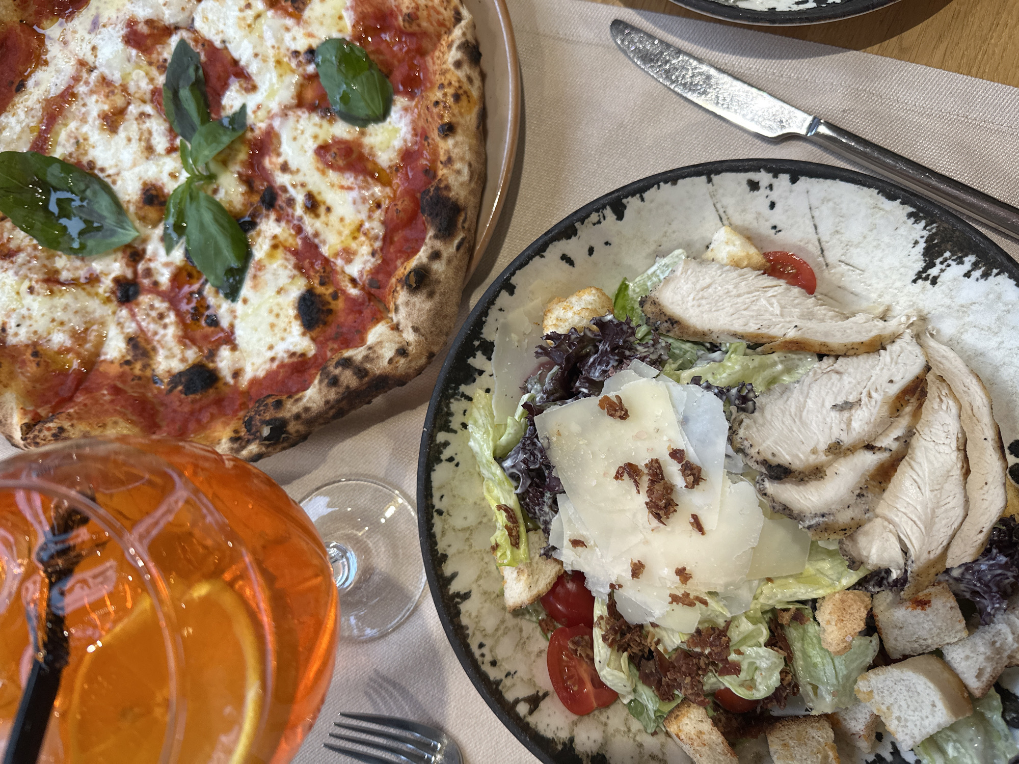 The scene is a close-up shot of a table setting, featuring a meal consisting of a pizza, a chicken Caesar salad, and an orange cocktail. The pizza has a thin crust with a charred edge, topped with tomato sauce, cheese, and basil leaves. The chicken Caesar salad is in a speckled ceramic bowl with a black rim and contains romaine lettuce, croutons, sliced chicken breast, cherry tomatoes, parmesan cheese, and bacon bits. A glass of orange cocktail is visible in the lower left, near a fork. A knife is placed on a beige tablecloth near the salad. The location is not discernible as Dobrich, Bulgaria, based on the provided image. There are no people visible.