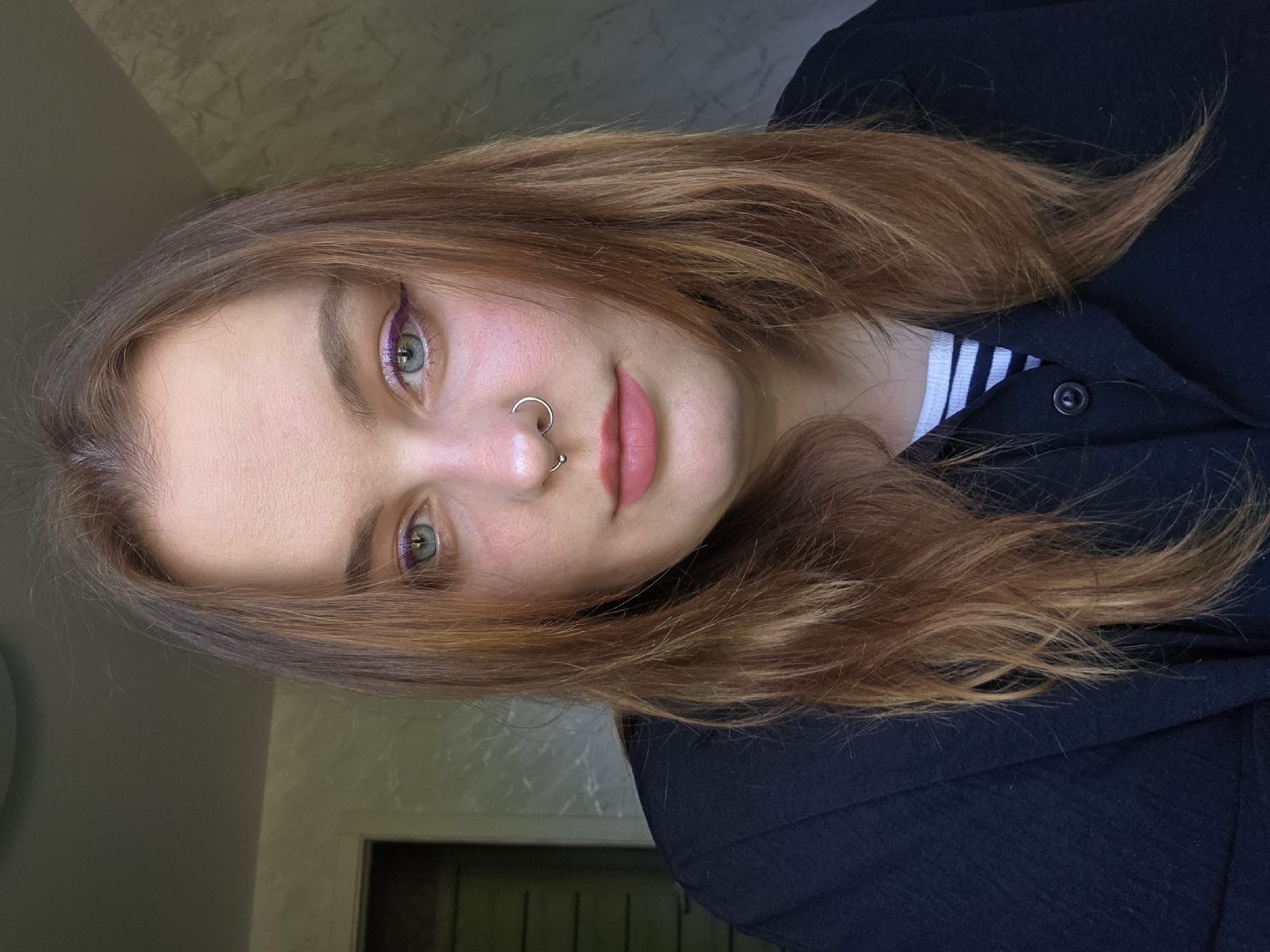 A medium shot shows a young woman with light green eyes and auburn hair, wearing a black jacket and a striped shirt underneath. She has a silver nose ring. Her makeup includes a purple eyeliner and light pink lipstick. The background is a plain, light gray wall and a glimpse of a dark door. The location is unknown.