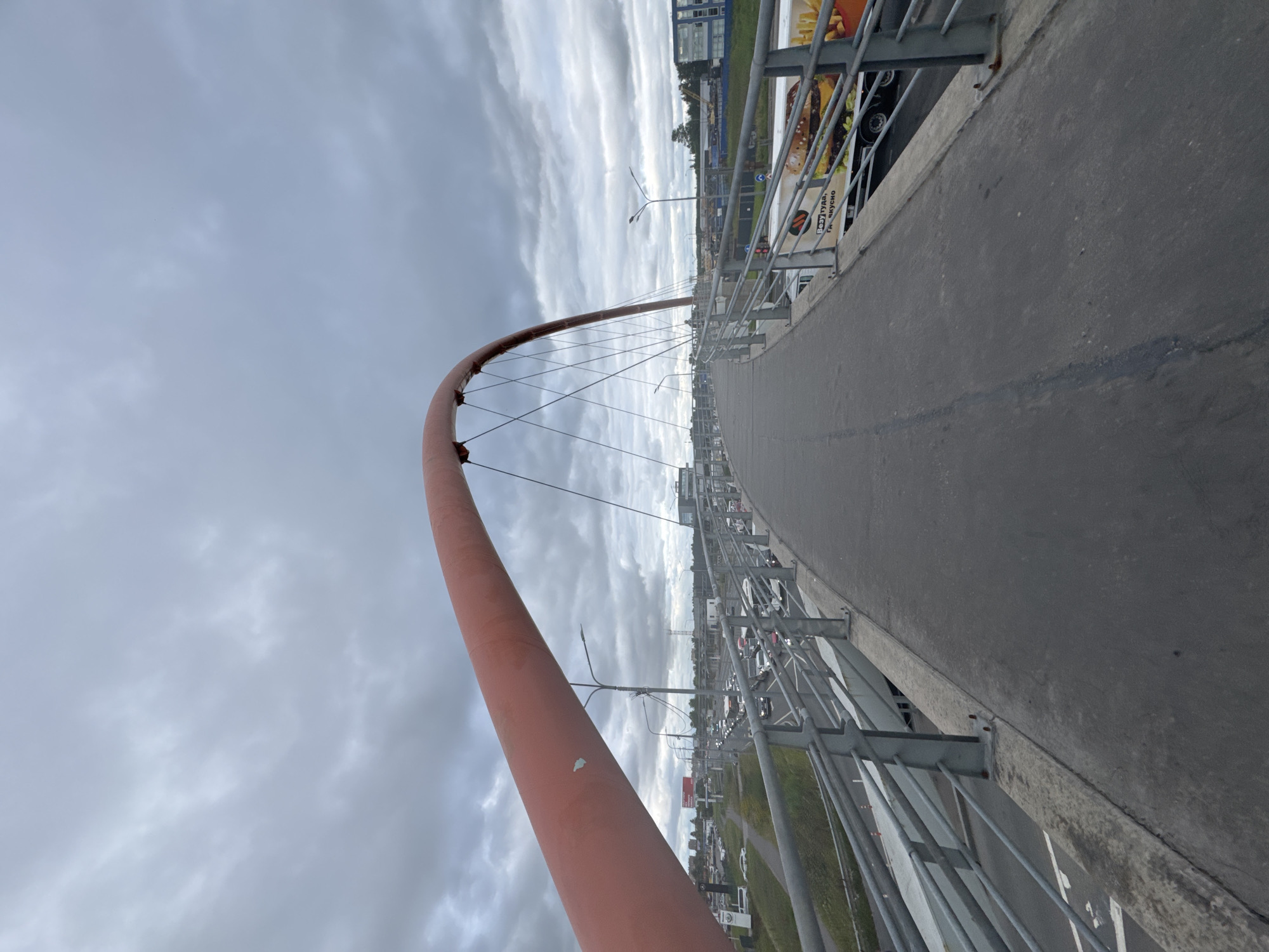 A pedestrian bridge with a red arch spans over a traffic-congested highway. The sky is overcast with gray clouds. The bridge deck is made of asphalt and has metal railings on both sides. A large truck with an advertisement for a fast-food restaurant is visible on the highway below. The location is likely Saint Petersburg, Russia, as suggested by the Cyrillic text on the truck's advertisement.