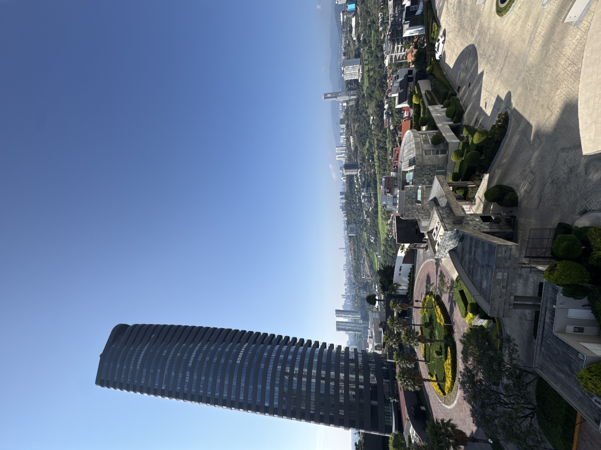 A view from a high vantage point in Huixquilucan de Degollado, Mexico. A tall, dark, modern skyscraper with a curved facade dominates the foreground. The city sprawls in the midground, displaying a mix of buildings, green spaces, and distant hills. To the right of the skyscraper, there is a circular driveway with a landscaped island in the center, featuring palm trees and flower beds. Adjacent to the driveway, there are buildings with sloped roofs. Above these structures, there is a residential area with homes and gardens. The sky is a clear, bright blue, occupying the upper portion of the frame.