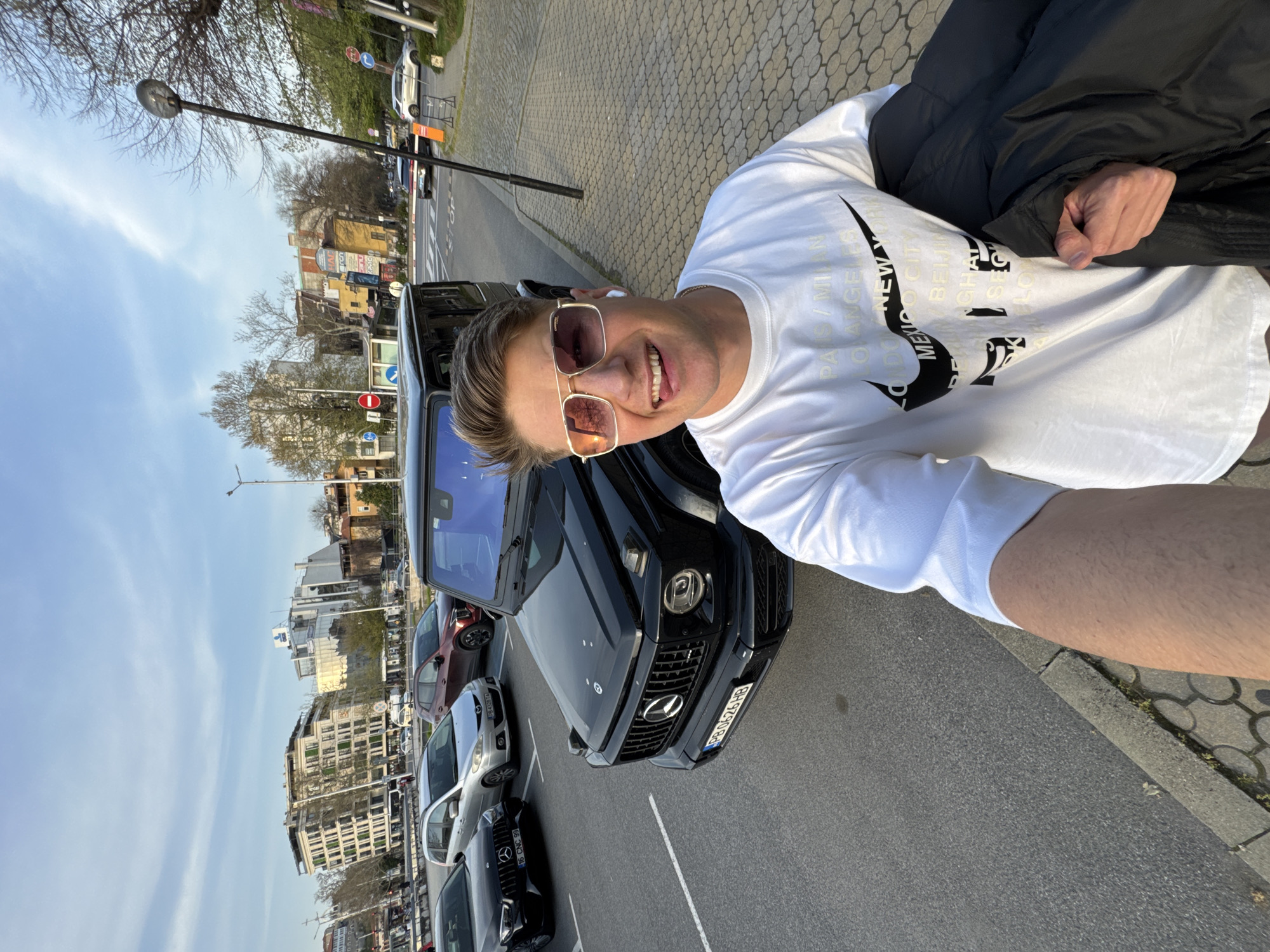A man with blond hair and wearing rose-tinted glasses poses for a selfie in front of a black Mercedes G-Wagon. He is wearing a white t-shirt with black Nike logos and lettering. A black coat is draped over his shoulder. Several parked cars are visible behind the G-Wagon. The scene appears to be a street in an urban setting, with buildings visible in the background. The license plate reads "PB 0626 HB" and "16 CNC 99." Based on the Cyrillic alphabet on the license plate and the style of the architecture, the location is likely Plovdiv, Bulgaria.