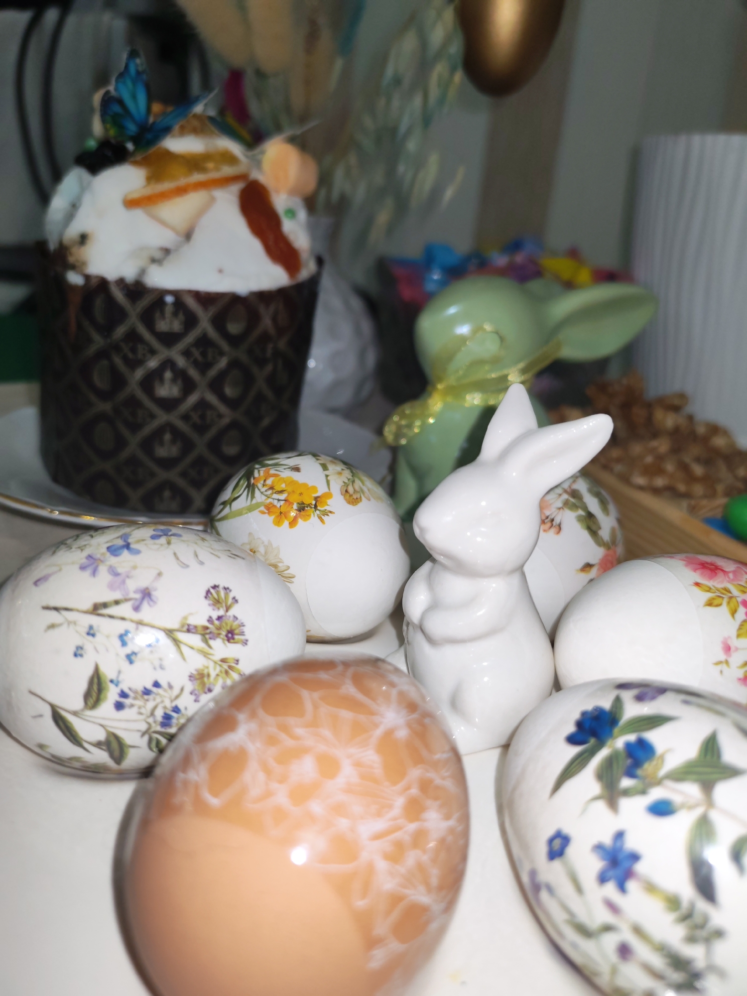 The image shows an Easter still life. A tall Easter cake ("kulich") decorated with white frosting, candied fruits, and a blue butterfly sits on a white plate. In front of the cake are several Easter eggs, some with floral decorations and one with a marbled brown pattern. A small white ceramic bunny stands among the eggs, and a green ceramic bunny with a golden ribbon around its neck is visible behind. In the background, there are various blurred objects, including what appear to be candies and walnuts. The location, Izhevsk, Russia, is not discernible from the image alone. The scene is indoors, likely on a table or countertop. There are no people visible in the image.