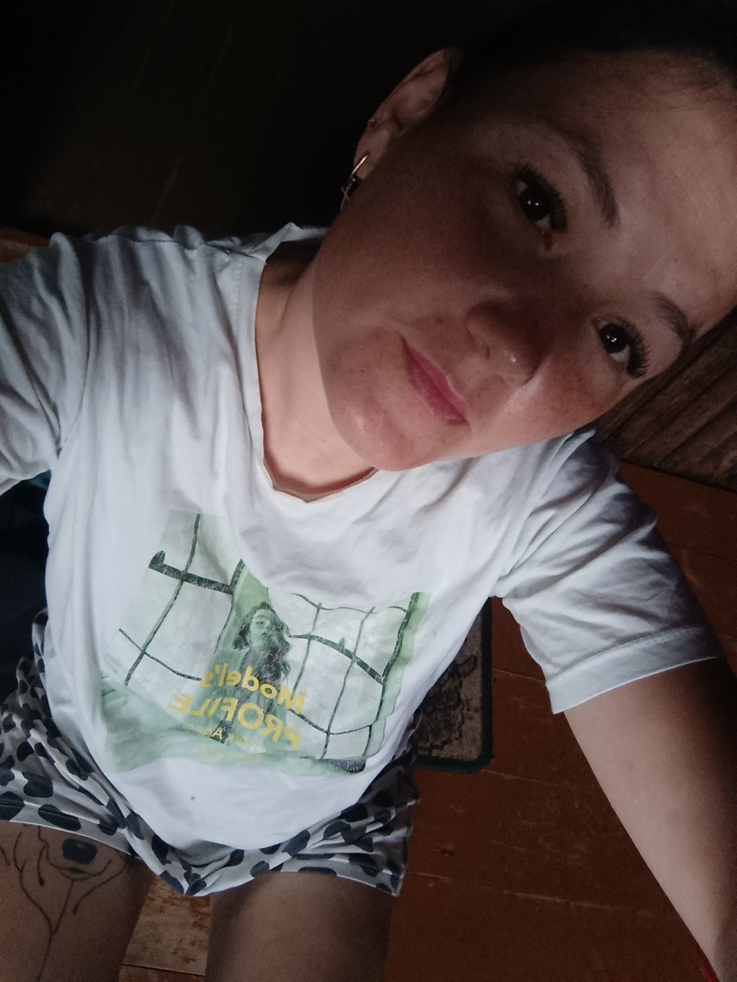 The image is a close-up, high-angle selfie of a young woman indoors. She has fair skin with freckles, dark eyebrows, dark eyelashes, and pink lipstick. Her hair is dark and pulled back. She is wearing a white t-shirt with a graphic print that includes the words "Model" and "Profile" backwards. She is also wearing patterned shorts. The background appears to be a wooden wall and floor. The lighting is dim, suggesting an indoor environment. There is no indication of the city of Karsha, Russia, or any specific location information.