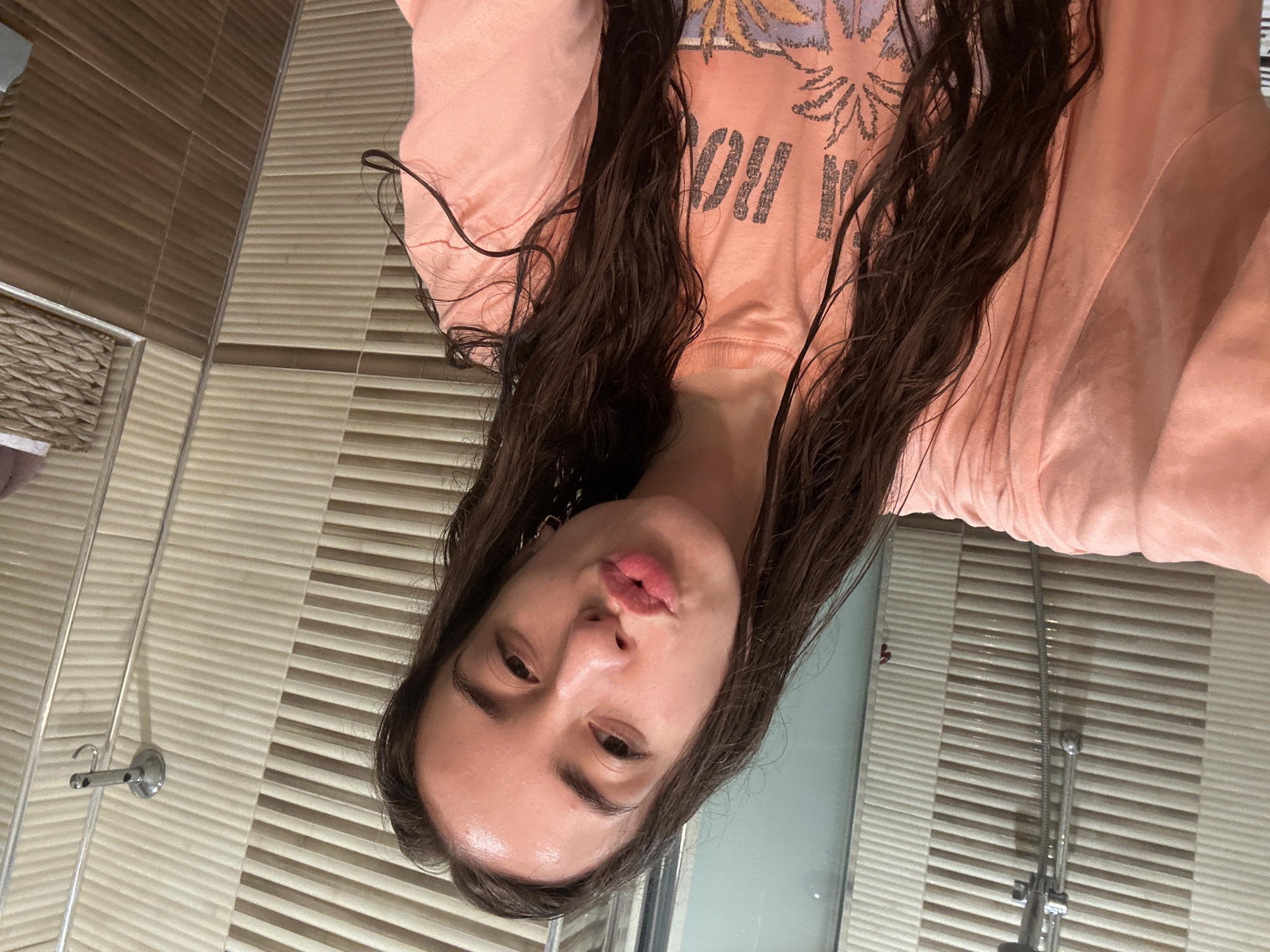 A young woman with long, dark, wet hair is posing for a selfie in a bathroom. She is wearing a peach-colored t-shirt with a graphic print. Her face is turned slightly to the side, and she is puckering her lips. The bathroom features tiled walls in shades of beige and brown. A shower head is visible on the left side of the image, and a wicker basket is on the right. The lighting suggests it is daytime, possibly after a shower. The image does not contain any recognizable landmarks or elements that would allow for its specific location within the city of Shumen, Bulgaria.