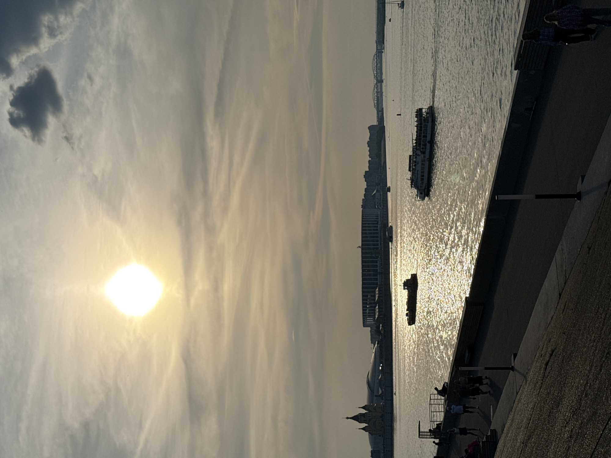 This image captures a sunset over the Volga River in Nizhny Novgorod, Russia. The sun, a bright orb, hangs in a sky filled with wispy clouds. In the foreground, a concrete embankment lines the river, with a pathway where several people are walking and standing. The river itself is calm, reflecting the sunlight and dotted with boats. In the distance, the cityscape of Nizhny Novgorod is visible, featuring the Strelka architectural ensemble with the Alexander Nevsky Cathedral and the Nizhny Novgorod Stadium. Bridges span the river in the far distance. The overall scene is one of a serene evening by the river.