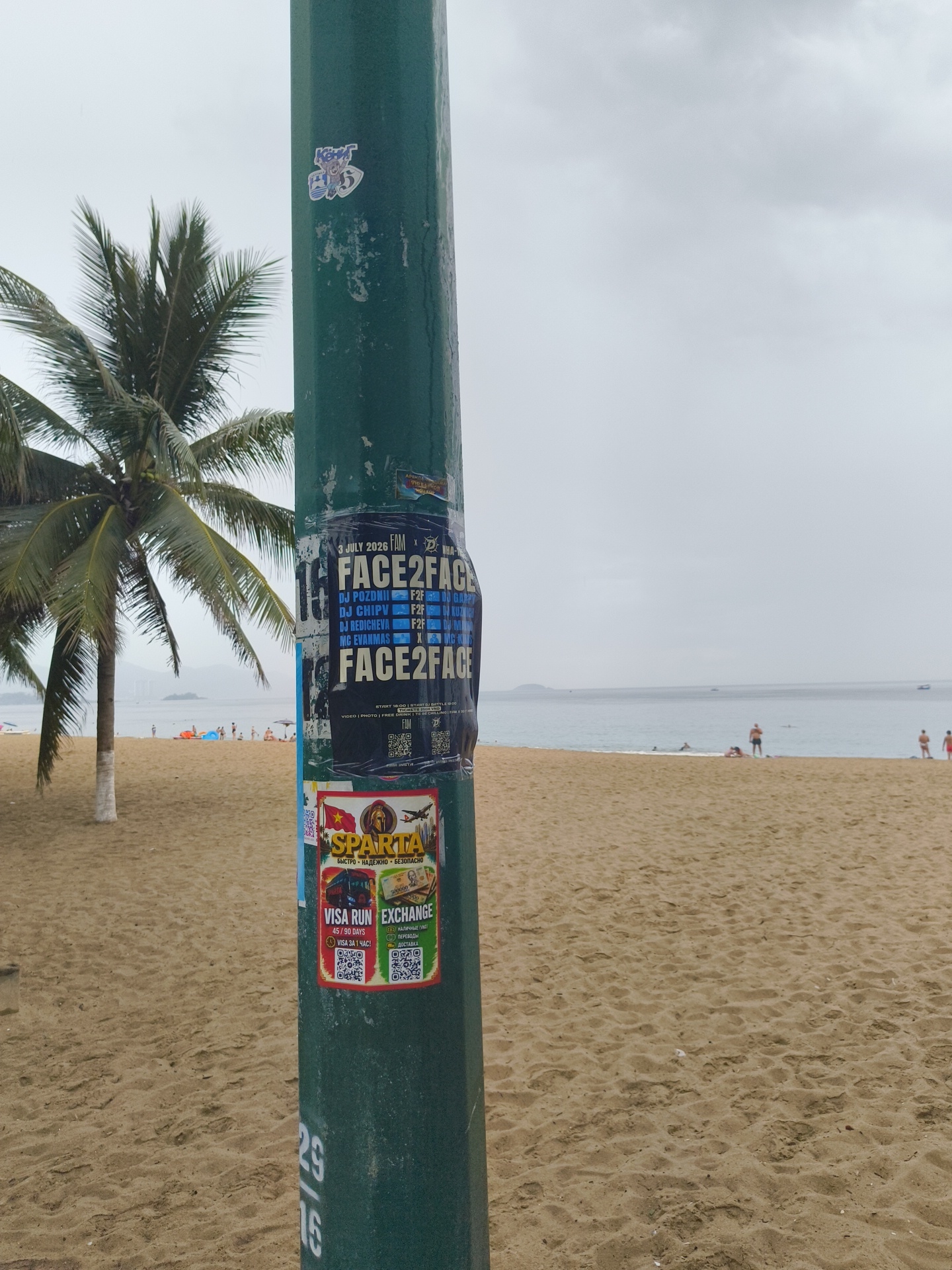 An overcast day at Nha Trang beach. A weathered green lamppost stands prominently in the foreground, adorned with multiple stickers and flyers. A large black flyer, titled "FACE2FACE," advertises an event on "3 July 2026" featuring several DJs and an MC. Below this, a red-bordered sticker for "SPARTA" promotes "FAST • RELIABLE • SAFE" services, along with a "VISA RUN EXCHANGE" sticker detailing services for "45 / 90 DAYS" and "VISA FOR 1 HOUR." Two QR codes are visible on the "VISA RUN EXCHANGE" sticker. To the left of the lamppost, a palm tree sways gently. In the background, the sandy beach stretches towards a calm, grey sea. Several people are scattered across the beach and in the water, some appearing to be sunbathing or swimming. In the distance, a few boats are visible on the horizon. The overall scene is one of a typical beach day, albeit under cloudy skies, with commercial advertisements integrated into the public space.