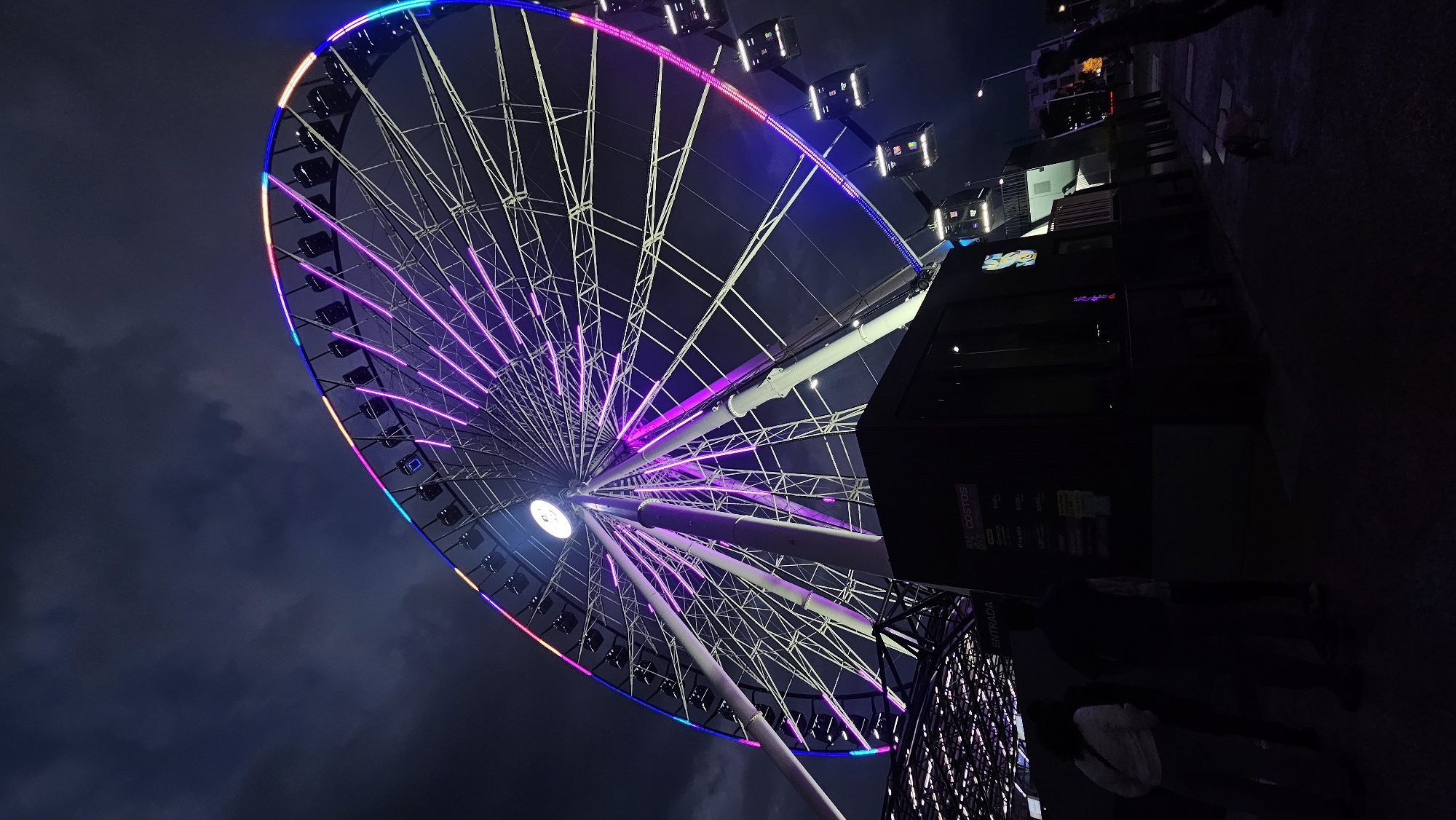 The image is a low-angle shot of a brightly lit Ferris wheel against a dark sky, likely at night. The Ferris wheel's rim is adorned with a sequence of colorful lights, transitioning through shades of blue, pink, and orange. Purple lights illuminate the spokes of the wheel, creating a radial pattern. The central hub of the wheel has a bright white light.
To the right of the Ferris wheel, there is a dark, rectangular structure that appears to be a building. The building has some signage and displays, but details are difficult to discern due to the darkness. In the foreground, there is part of a metal fence or barrier. Silhouetted figures of people can be seen near the base of the building and at the bottom of the frame.
Given the architecture and available information, this is the "Estrella de Puebla" (Puebla Star) in Puebla, Mexico. The environment appears to be an urban setting with a cloudy or dark sky providing contrast to the bright lights of the Ferris wheel.